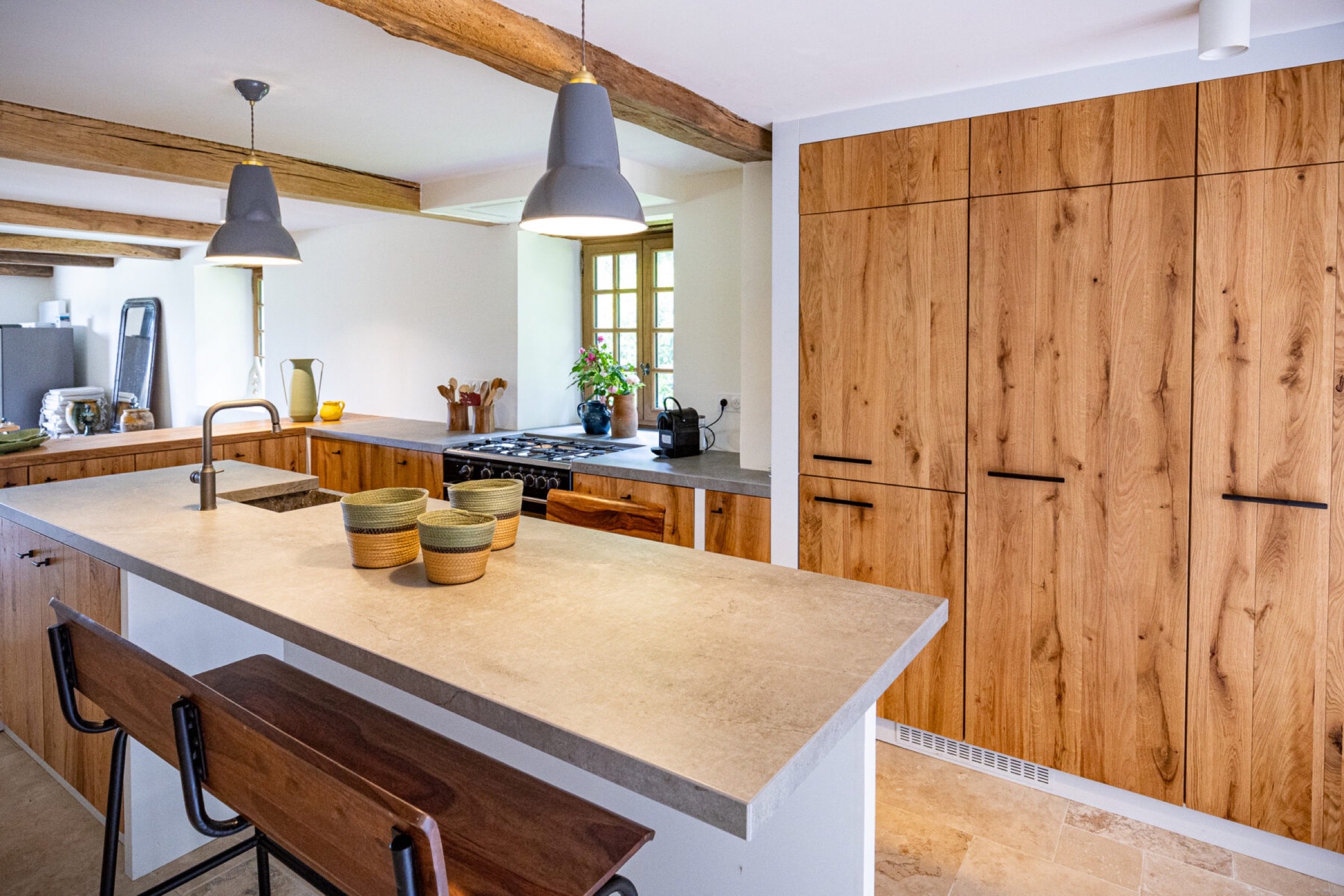 Une cuisine moderne dot&eacute;e d'un &icirc;lot de couleur claire avec un &eacute;vier et un robinet int&eacute;gr&eacute;s, associ&eacute; &agrave; deux tabourets de bar en bois. La cuisine comprend des armoires en bois, des comptoirs en pierre et deux suspensions suspendues au-dessus de l'&icirc;lot. Une cuisini&egrave;re &agrave; gaz avec hotte est situ&eacute;e contre le mur.