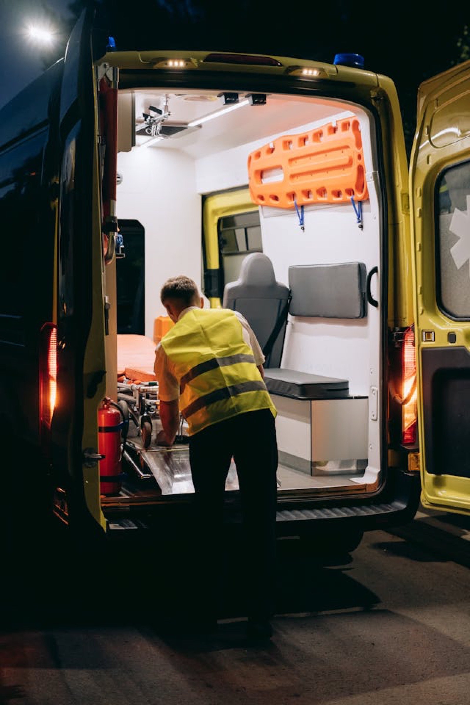 Une personne portant un gilet r&eacute;fl&eacute;chissant charge ou d&eacute;charge un brancard &agrave; l'arri&egrave;re d'une ambulance jaune - dot&eacute;e d'un am&eacute;nagement de camion moderne - de nuit dans le Nord de Tarbes. Du mat&eacute;riel m&eacute;dical et une planche dorsale sont visibles &agrave; l'int&eacute;rieur.