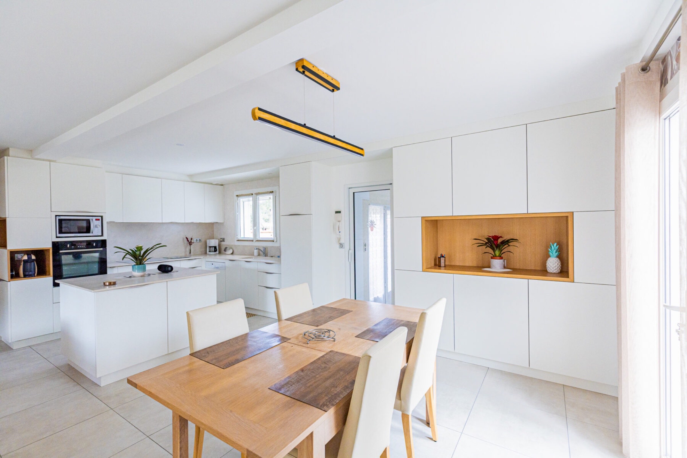 Cuisine moderne avec armoires blanches, table &agrave; manger en bois, &eacute;lectrom&eacute;nagers encastr&eacute;s et plantes d&eacute;coratives sous un &eacute;clairage naturel et artificiel, avec un am&eacute;nagement personnalis&eacute; de l'Agencement sur mesure Tarbes.