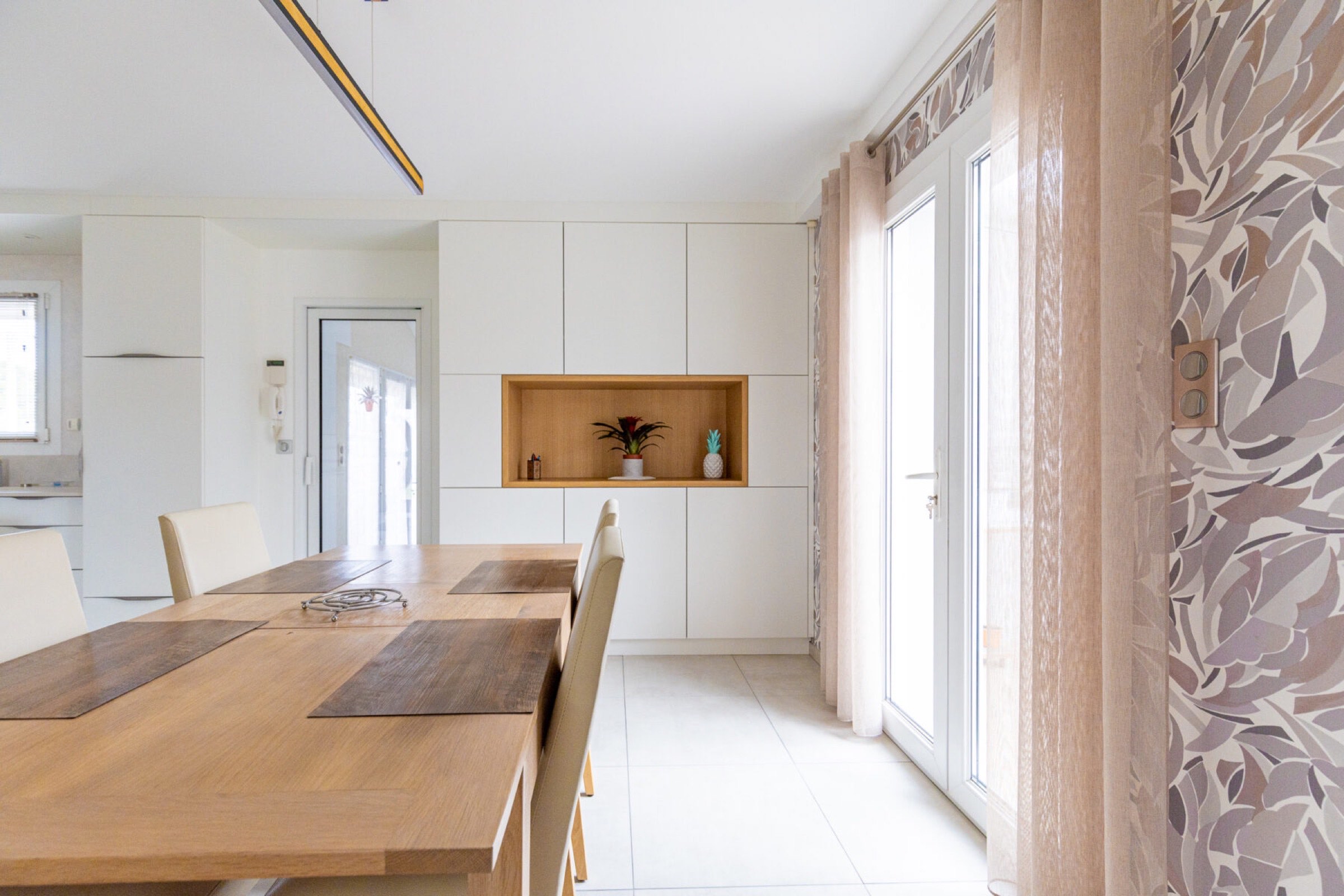 Une cuisine lumineuse et moderne avec des murs blancs, une grande table &agrave; manger en bois et des armoires encastr&eacute;es con&ccedil;ues pour la s&eacute;r&eacute;nit&eacute;. La lumi&egrave;re naturelle p&eacute;n&egrave;tre &agrave; travers une porte vitr&eacute;e et le d&eacute;cor comprend un minimum de