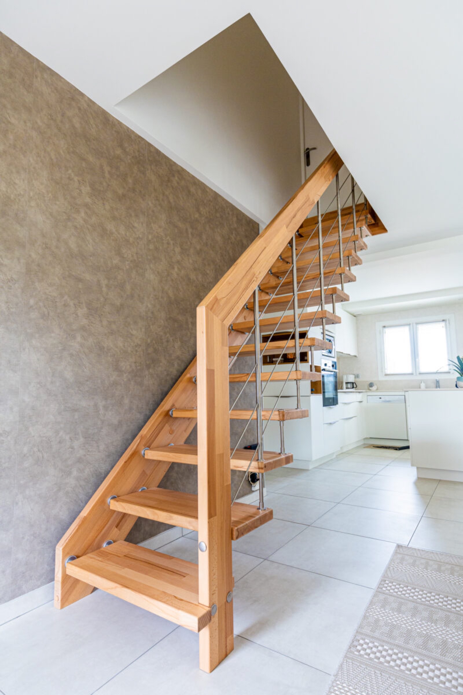 Un int&eacute;rieur moderne comprenant un escalier en bois avec des contremarches ouvertes et des garde-corps en m&eacute;tal, montant &agrave; c&ocirc;t&eacute; d'un mur gris textur&eacute;, menant &agrave; un &eacute;tage sup&eacute;rieur visible en arri&egrave;re-plan apr&egrave;s une r&eacute;novation de maison.