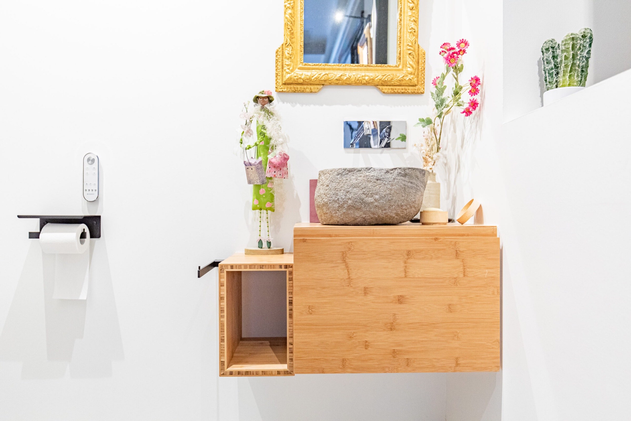 Un coin salle de bain moderne et compact comprenant une vanit&eacute; en bois clair avec une &eacute;tag&egrave;re ouverte, un lavabo en pierre, compl&eacute;t&eacute; par des fleurs color&eacute;es dans des pots et un miroir au cadre dor&eacute; sur le mur.