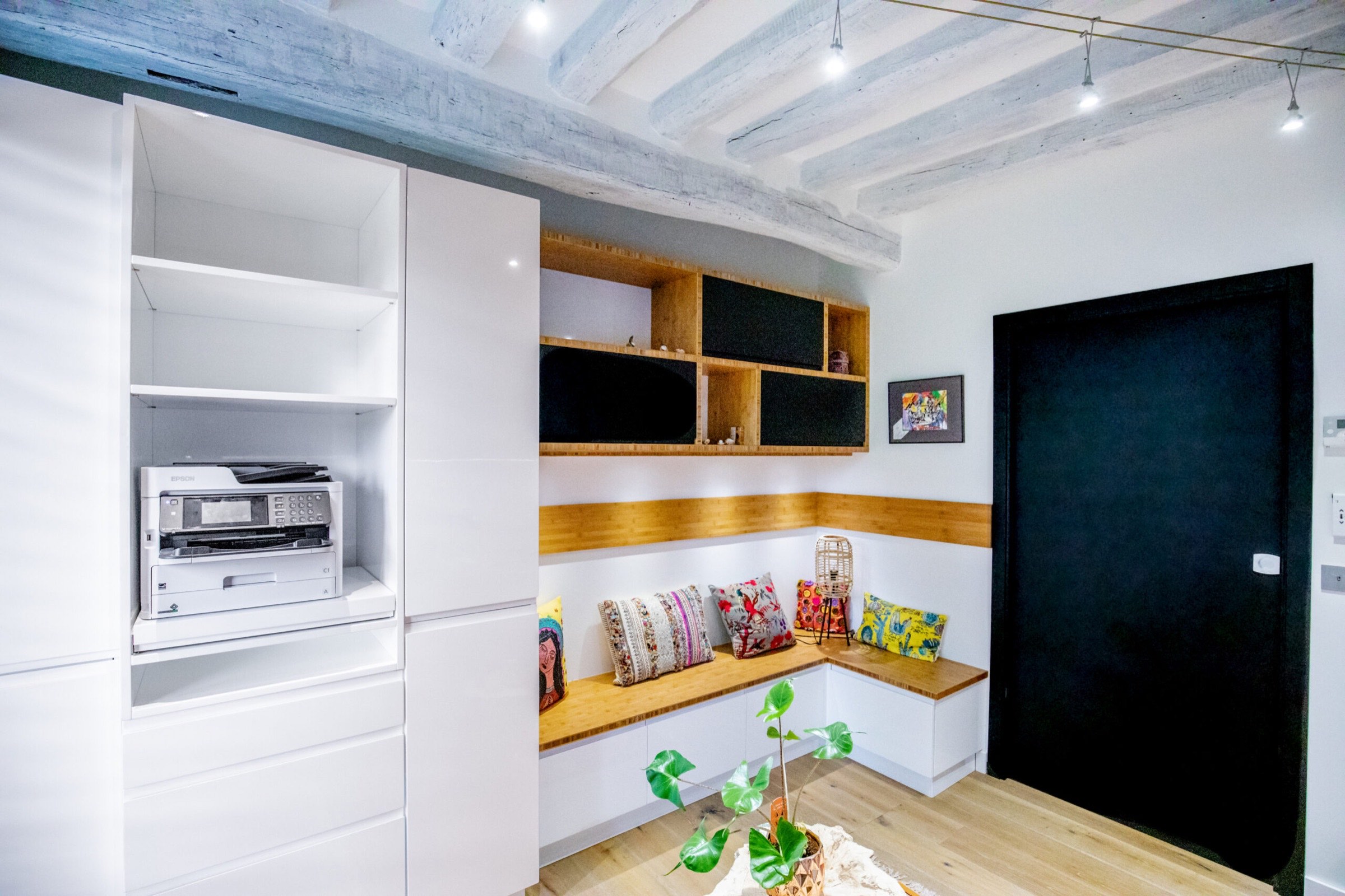 Coin cuisine moderne avec armoires blanches, micro-ondes int&eacute;gr&eacute;, un confortable banc en bois avec des coussins color&eacute;s et une plante d&eacute;corative, le tout sous un plafond de style industriel avec poutres apparentes et d&eacute;coration en bois.