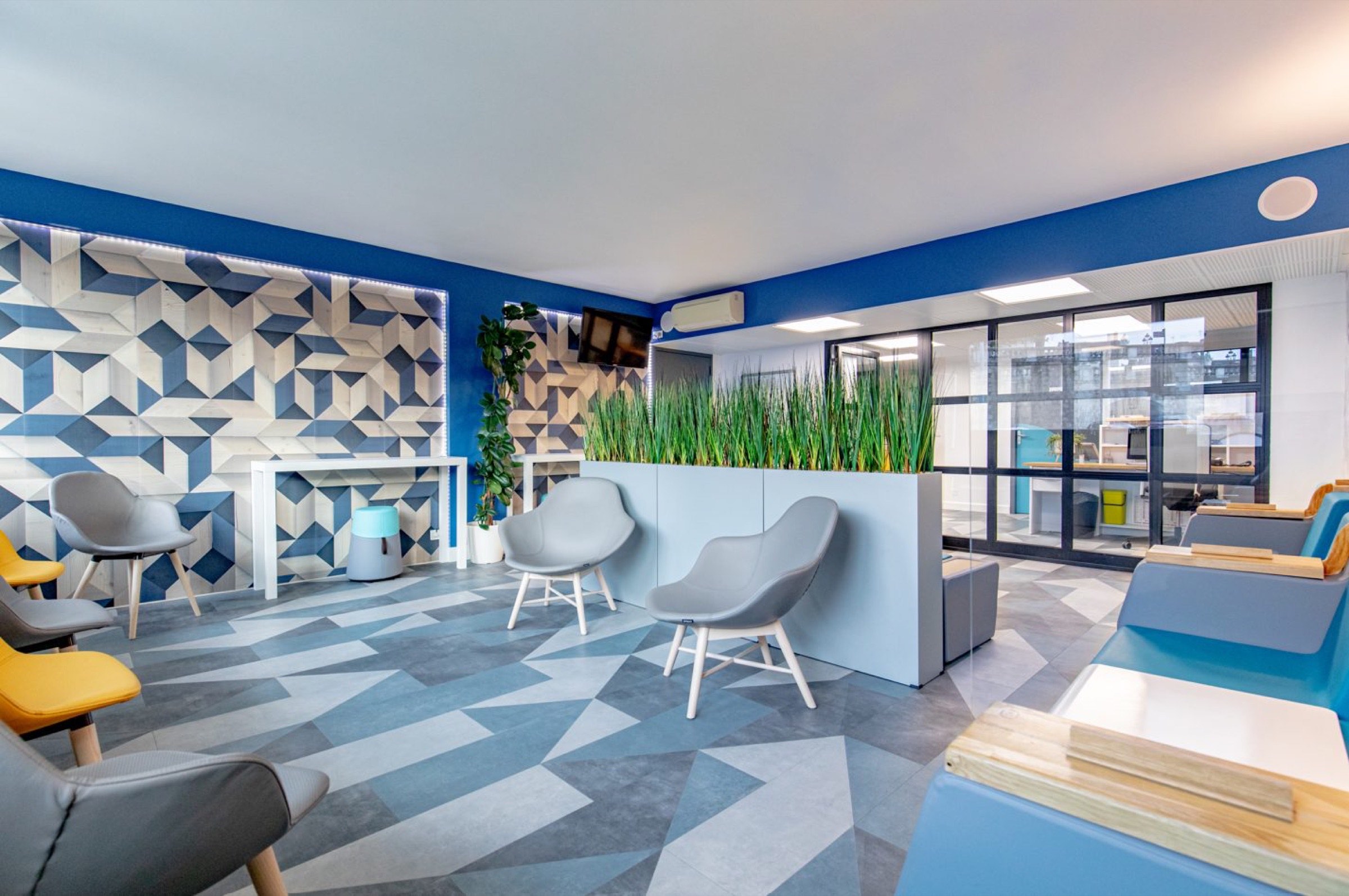 Un salon de bureau moderne et color&eacute; avec des &eacute;l&eacute;ments de design g&eacute;om&eacute;trique, con&ccedil;u par un architecte d'int&eacute;rieur &agrave; Tarbes, comprenant des coins salons confortables et des plantes d&eacute;coratives, favorisant un environnement de travail dynamique et cr&eacute;atif.