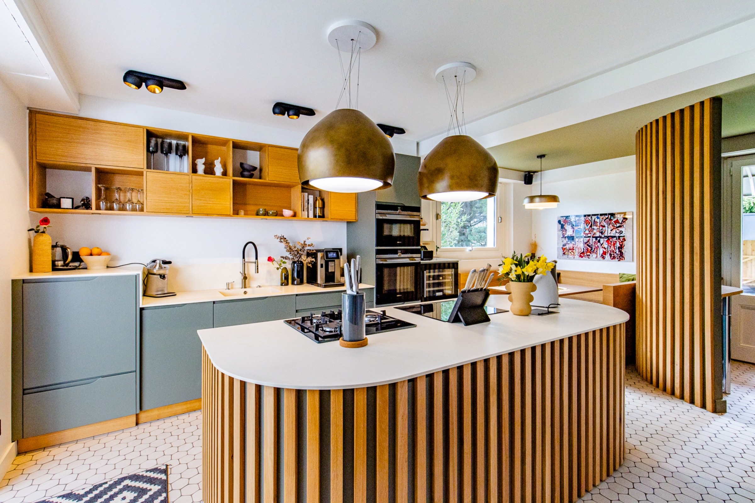 Une cuisine moderne et élégante comprenant un îlot central avec une table de cuisson, flanqué d'élégantes armoires en bois et de suspensions audacieuses en laiton. La pièce est agrémentée d'un dosseret à motifs