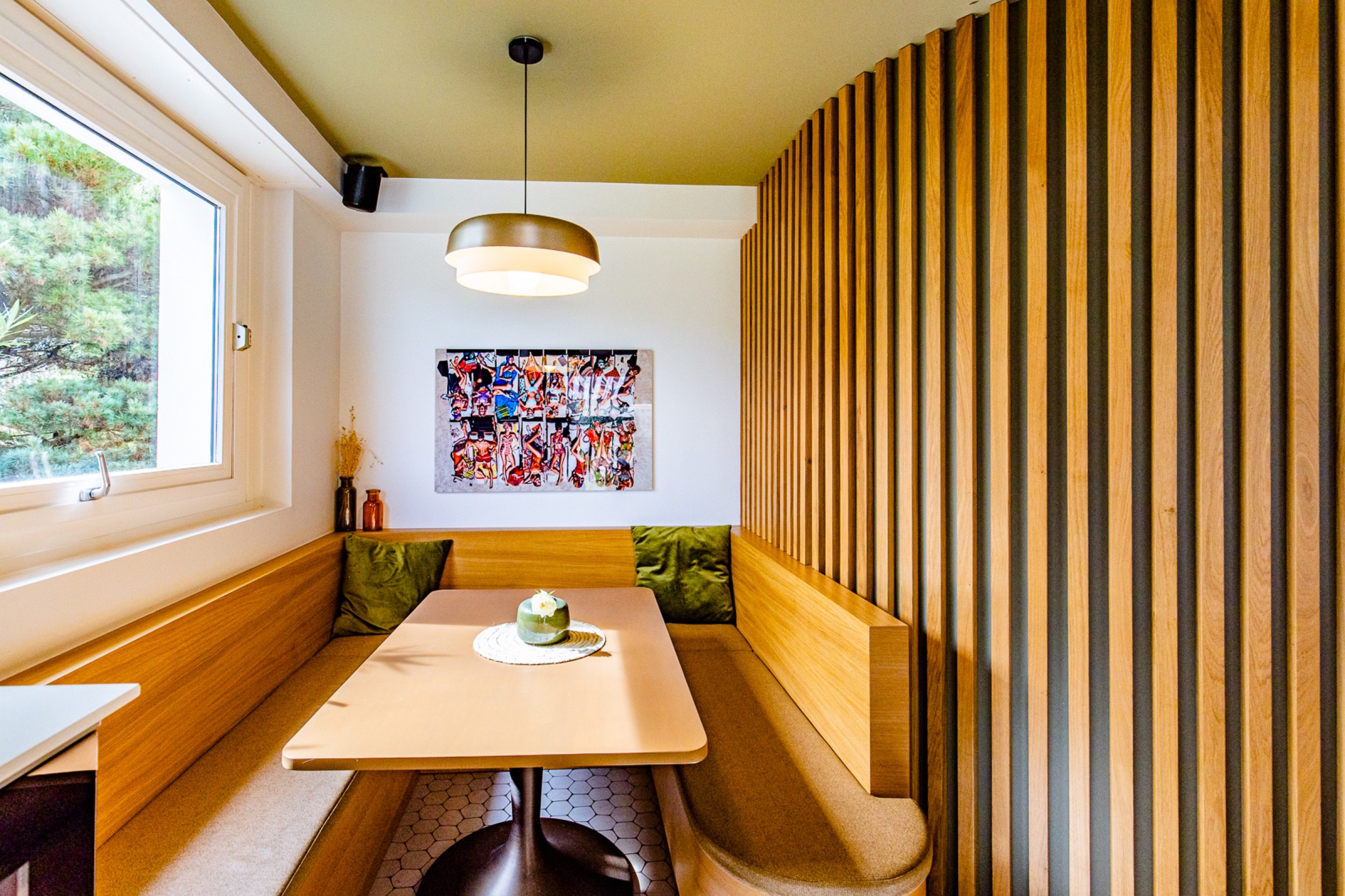 Un coin confortable avec un banc de salle à manger moderne, doté de coussins vert olive et de boiseries, accentué par une œuvre d'art murale vibrante sous un éclairage suspendu chaleureux. Un seul dessert repose sur la table,