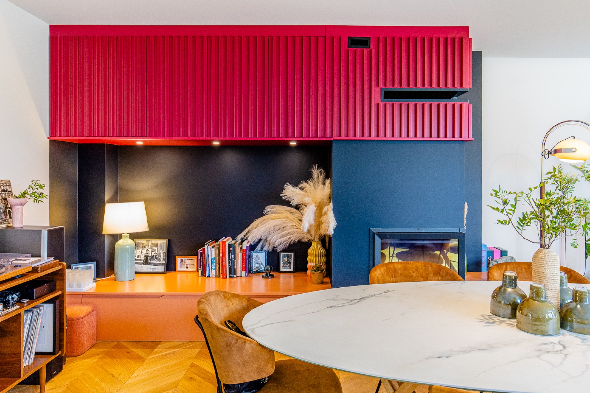 Une salle à manger moderne et élégante comprenant une table en marbre, des sièges confortables, un mélange d'armoires rouges vifs et un mur d'accent bleu profond, complétés par des éléments décoratifs et des plantes d'intérieur, tout en conservant