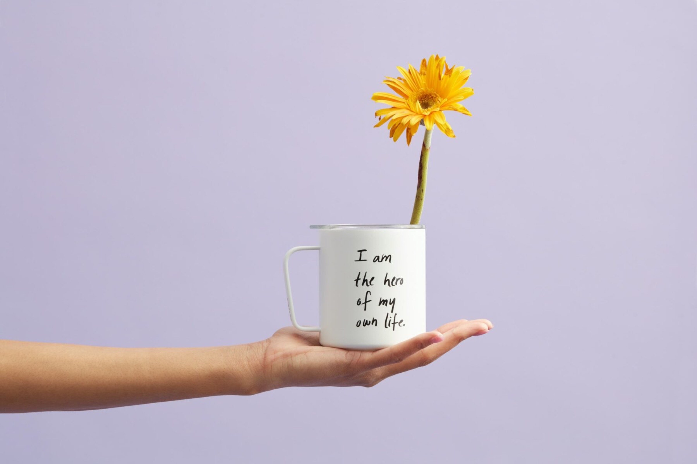Une main tenant une tasse blanche avec la citation inspirante "Je suis l'architecte d'int&eacute;rieur &agrave; Tarbes de ma propre vie" imprim&eacute;e dessus, avec une gerbera jaune vif