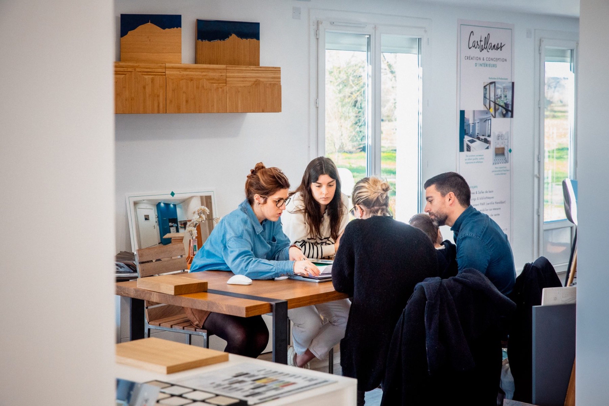 Quatre personnes sont assises autour d'une table dans un bureau lumineux, collaborant sur un projet de RSE. Des documents et des &eacute;chantillons sont &eacute;tal&eacute;s, tandis que la lumi&egrave;re du soleil filtre &agrave; travers de grandes fen&ecirc;tres en arri&egrave;re-plan, cr&eacute;ant un espace inspirant pour l'&eacute;quipe de Castellanos.