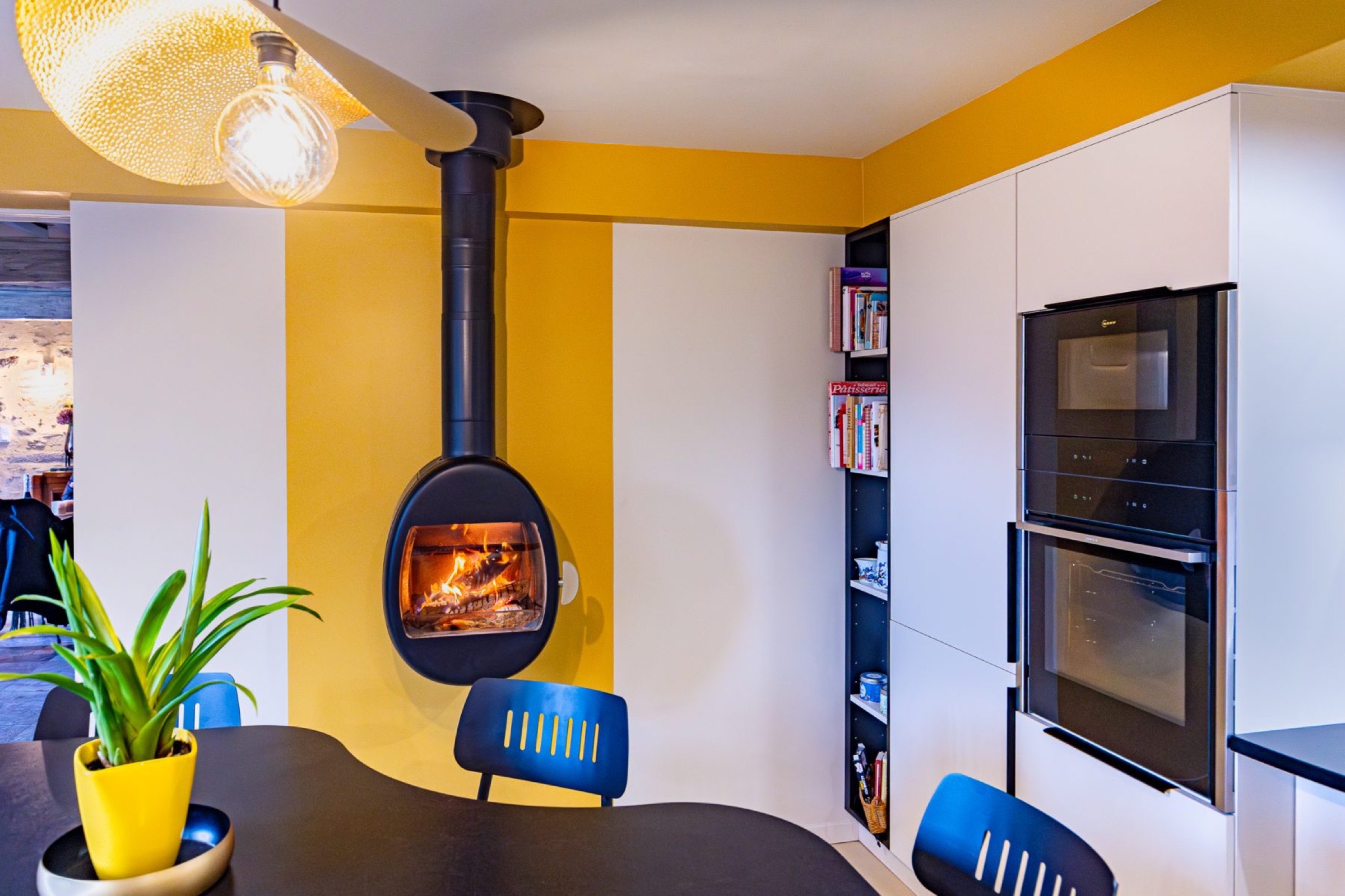 Cette cuisine moderne pr&eacute;sente des &eacute;l&eacute;ments de relooking avec des murs jaune vif et une chemin&eacute;e noire suspendue. Une table ronde avec une plante en pot et des chaises orne le premier plan, tandis que des armoires blanches encastr&eacute;es et un four mural se trouvent sur la droite. Une lampe sph&eacute;rique est &eacute;l&eacute;gamment suspendue au plafond.