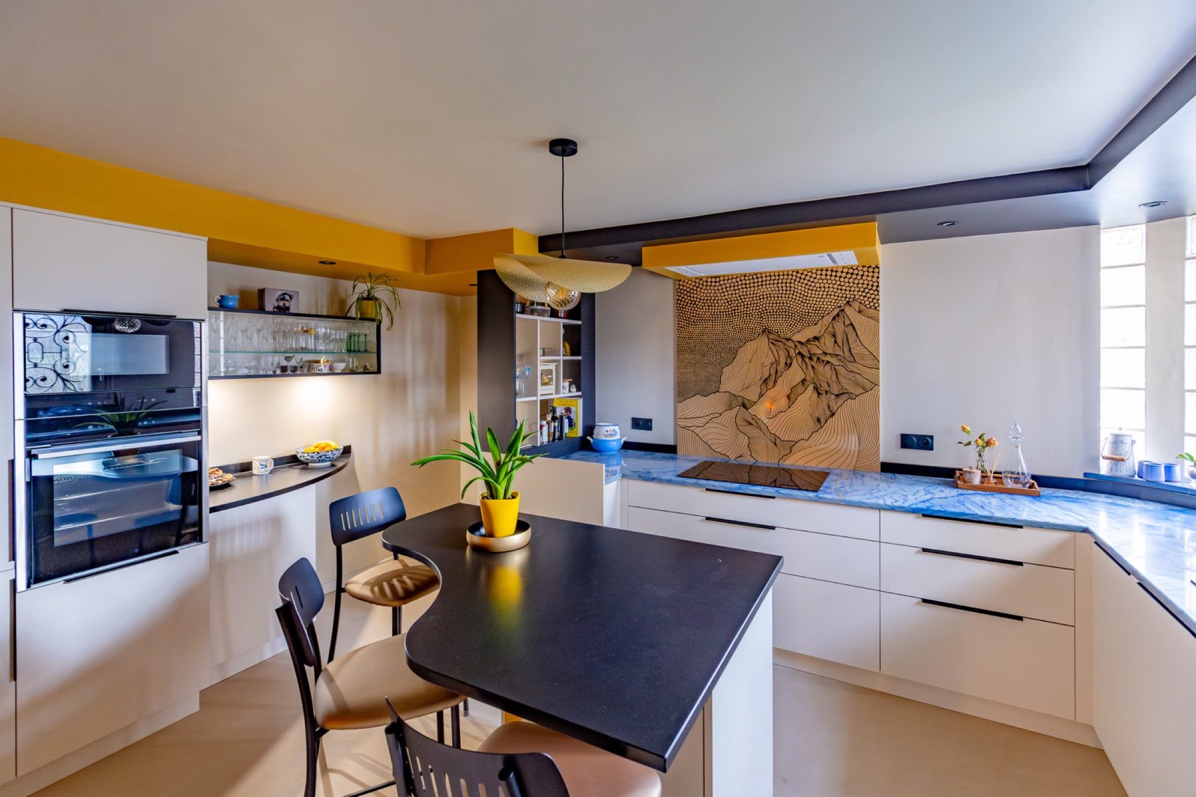Cette cuisine moderne, avec sa palette de couleurs jaune et blanche, offre un relooking simple. Elle comprend un &icirc;lot avec un comptoir noir &eacute;l&eacute;gant, des chaises et une plante en pot. Le dosseret au design montagnard ajoute du style tandis que les &eacute;tag&egrave;res pr&eacute;sentent la vaisselle et la d&eacute;coration. Un &eacute;clairage lumineux la rend accueillante mais suffisamment &eacute;l&eacute;gante.