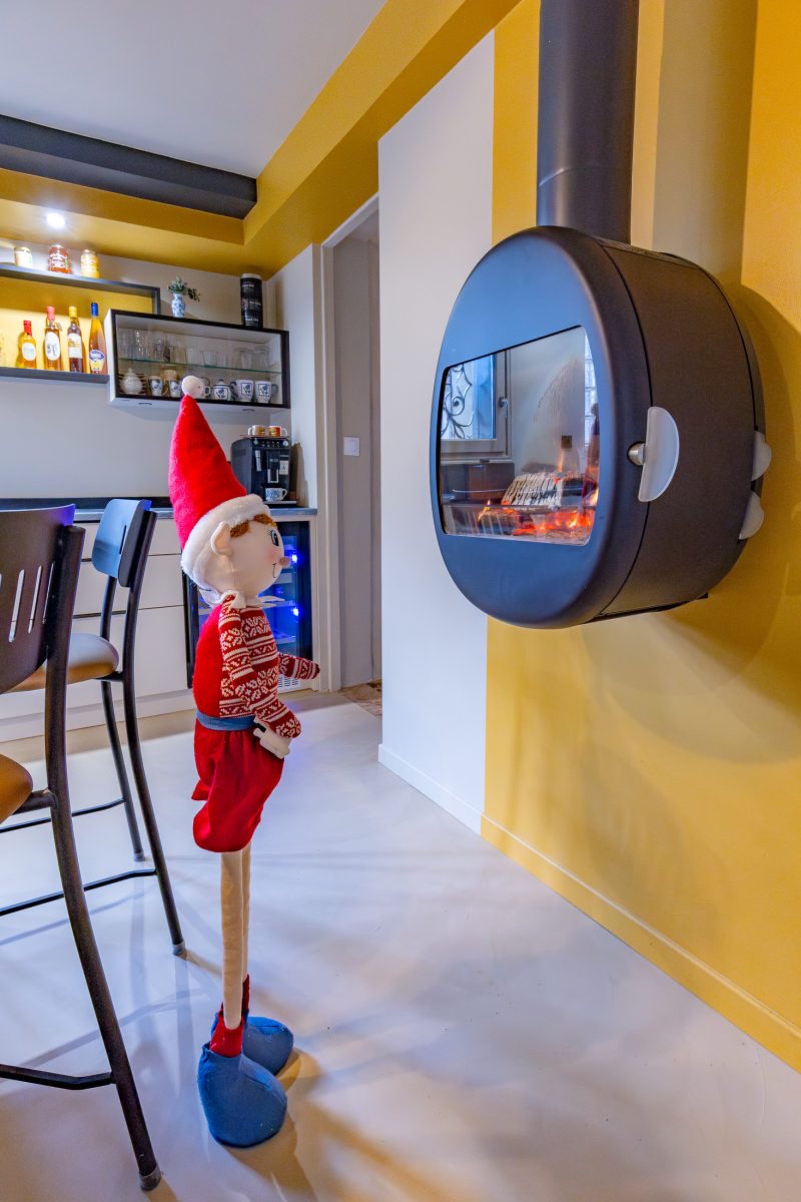 Une poup&eacute;e elfe festive, v&ecirc;tue d'une tenue rouge et de chaussures bleues, se tient debout face &agrave; une chemin&eacute;e murale moderne. La pi&egrave;ce pr&eacute;sente des murs jaunes et un bar avec des &eacute;tag&egrave;res de bouteilles, sugg&eacute;rant un relooking simple qui ajoute du caract&egrave;re &agrave; l'espace.