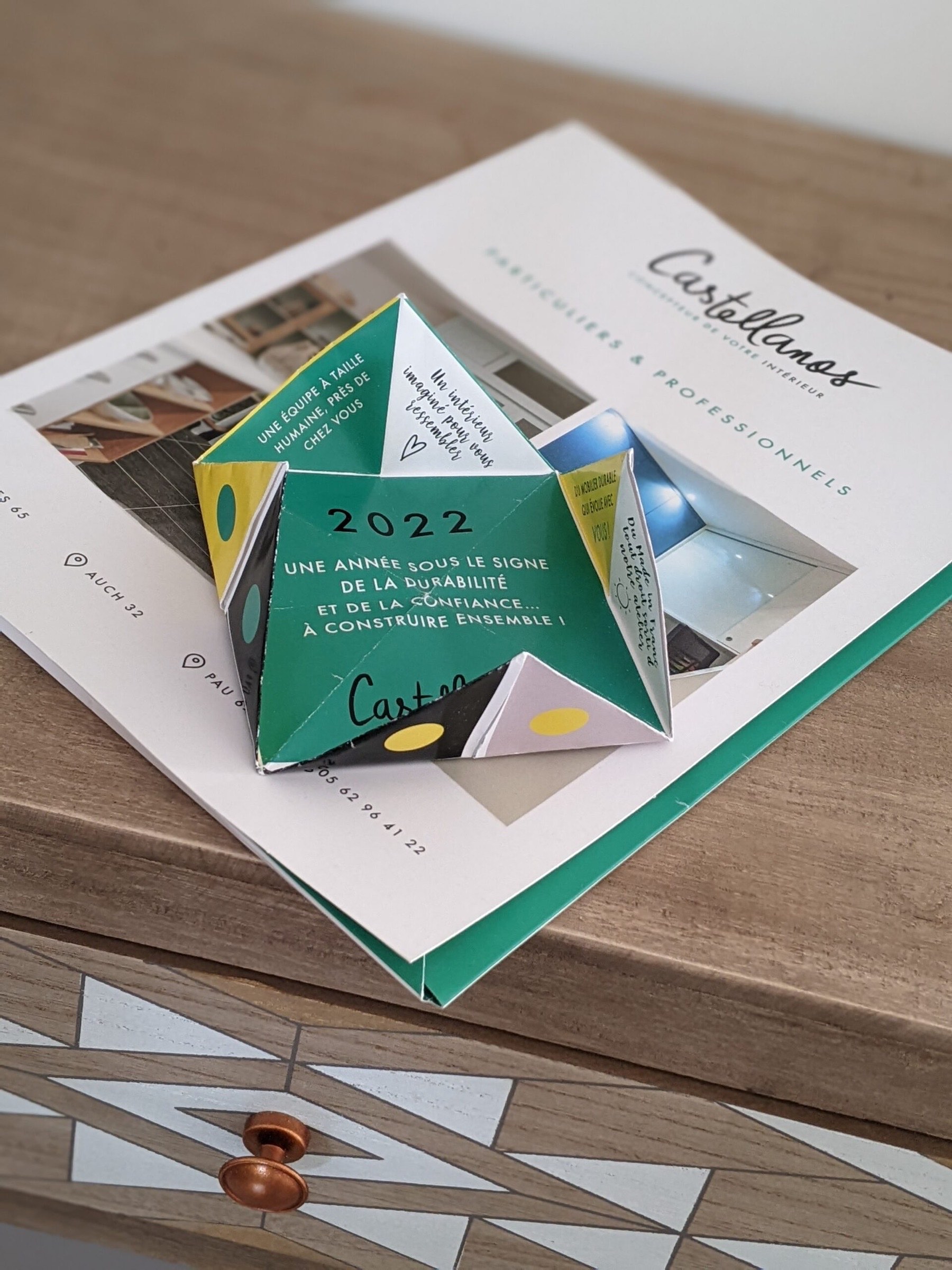 Une diseuse de bonne aventure en papier cr&eacute;ative reposant sur une brochure ouverte, pr&eacute;sentant un design graphique &eacute;l&eacute;gant et l'ann&eacute;e 2022 mise en &eacute;vidence, illustrant un support marketing ludique et interactif con&ccedil;u pour construire un ensemble.