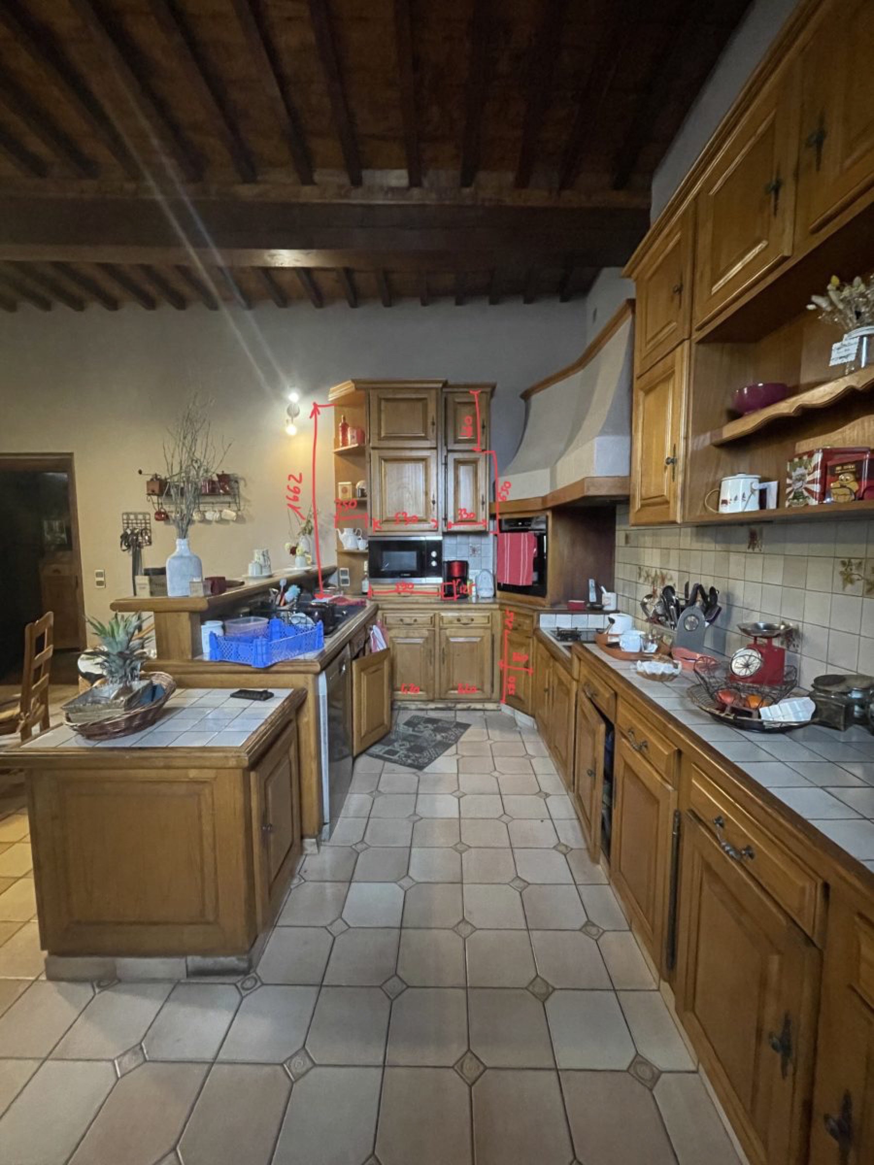 Une cuisine rustique avec des armoires en bois et des comptoirs carrel&eacute;s. Un &icirc;lot central accueille un panier, mettant en valeur un relooking simple. Les appareils &eacute;lectrom&eacute;nagers et les ustensiles de cuisine sont visibles, avec des marquages rouges et des num&eacute;ros sur l'armoire du fond. Les objets d&eacute;coratifs sont pos&eacute;s en toute confiance sur les &eacute;tag&egrave;res.