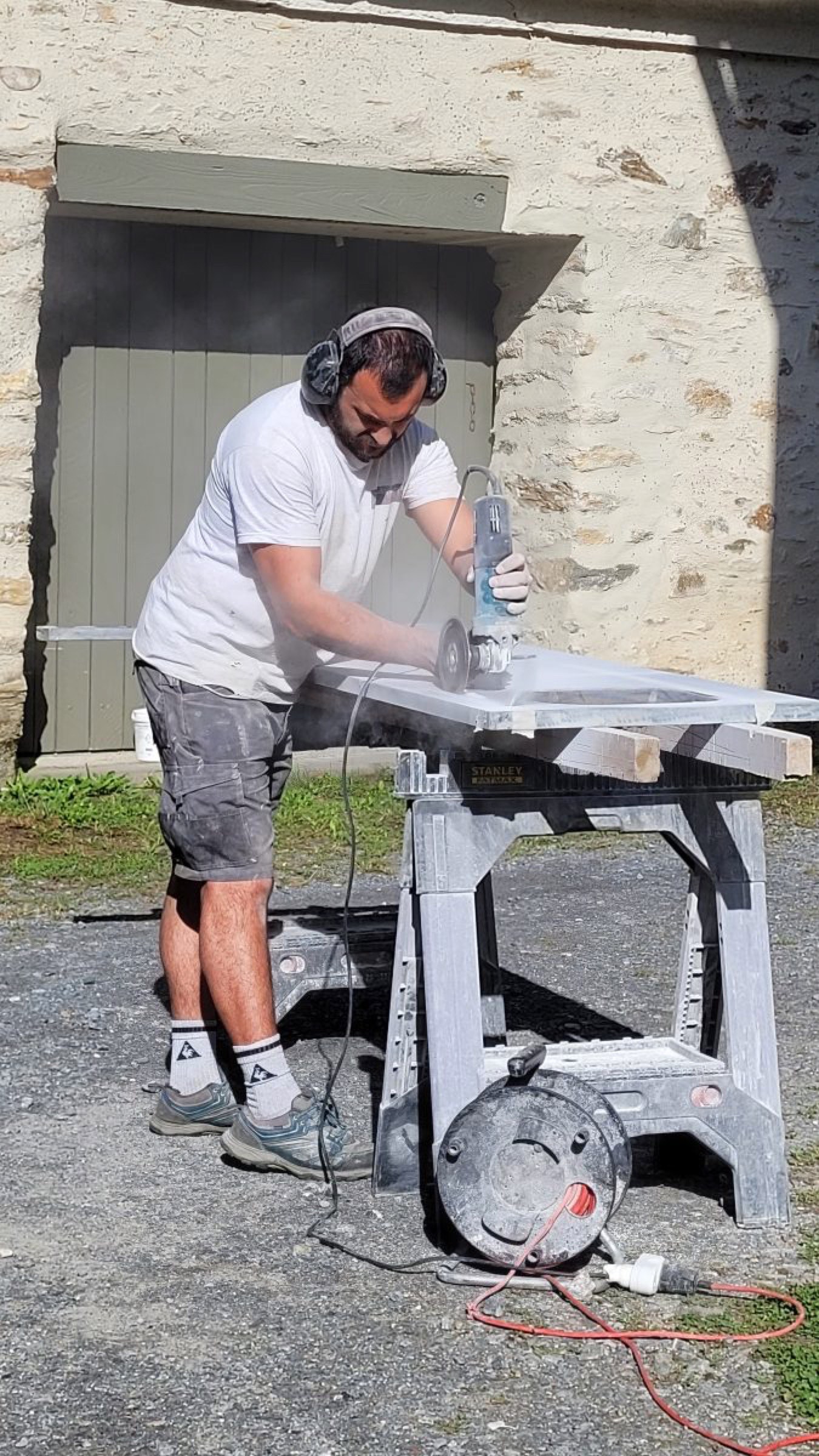 Un homme portant des &eacute;couteurs et des lunettes de s&eacute;curit&eacute; utilise une scie &eacute;lectrique sur un morceau de mat&eacute;riau fix&eacute; &agrave; un &eacute;tabli &agrave; l'ext&eacute;rieur. La poussi&egrave;re emplit l'air alors qu'il travaille sur son simple projet de r&eacute;novation par une journ&eacute;e ensoleill&eacute;e, avec un b&acirc;timent en pierre et une porte verte en arri&egrave;re-plan.
