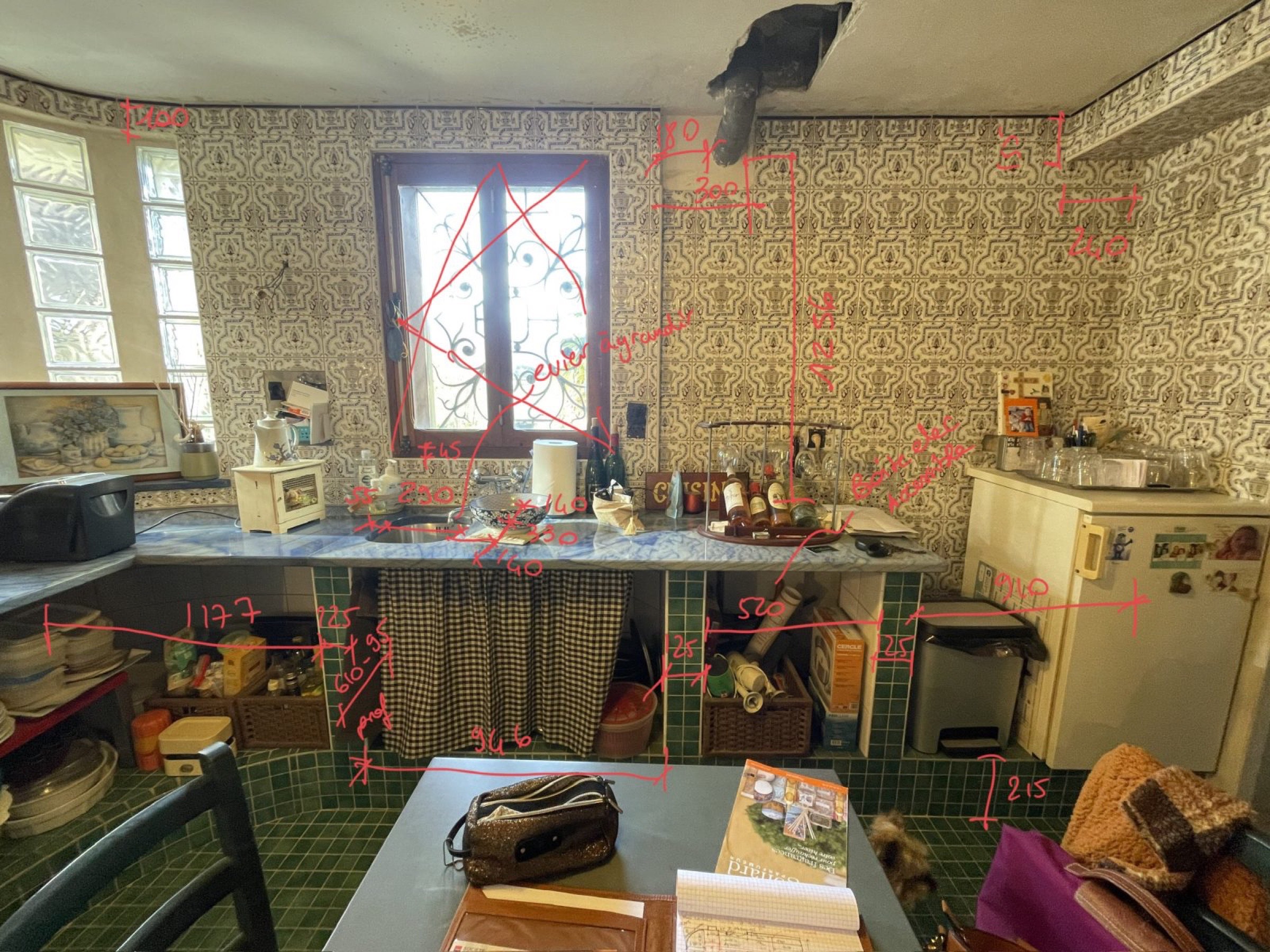 Une cuisine encombr&eacute;e avec du papier peint &agrave; motifs et un sol en carrelage vert. Le plan de travail contient divers objets tels que des bouteilles et des r&eacute;cipients. Des touches subtiles de relooking de la cuisine sont &eacute;videntes, renfor&ccedil;ant le charme. Des mesures en rouge sont dessin&eacute;es sur l'image. Une grande fen&ecirc;tre avec une grille d&eacute;corative fait face &agrave; l'ext&eacute;rieur.