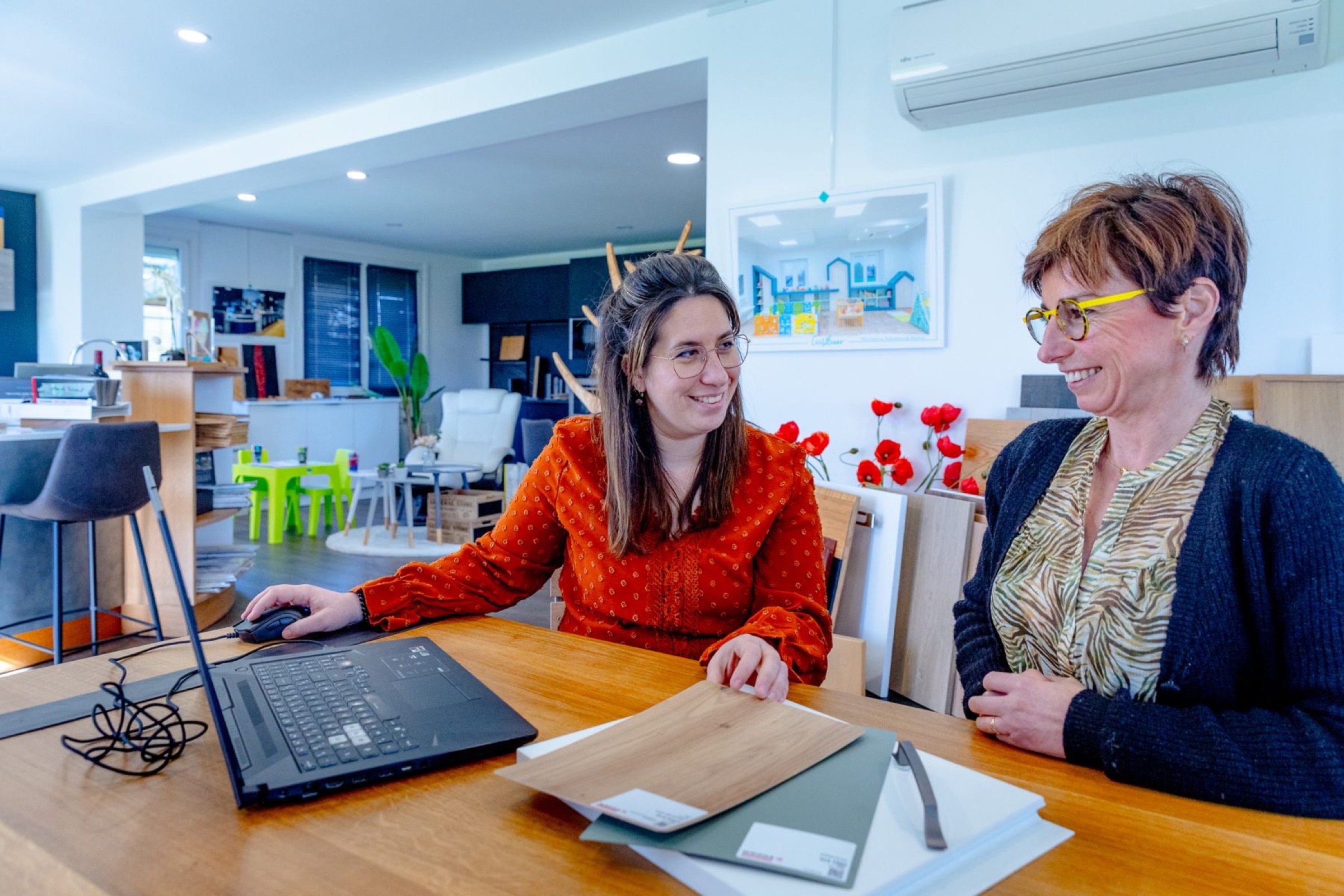 Deux femmes sont assises &agrave; une table en bois dans un bureau moderne et discutent de d&eacute;coration int&eacute;rieure. L'une d'elles, qui se concentre sur la r&eacute;novation de la cuisine, tient un &eacute;chantillon de bois, l'autre tape sur un ordinateur portable. Des papiers et d'autres mat&eacute;riaux sont dispos&eacute;s autour d'elles dans une pi&egrave;ce suffisamment lumineuse avec des &oelig;uvres d'art et des meubles color&eacute;s en arri&egrave;re-plan.