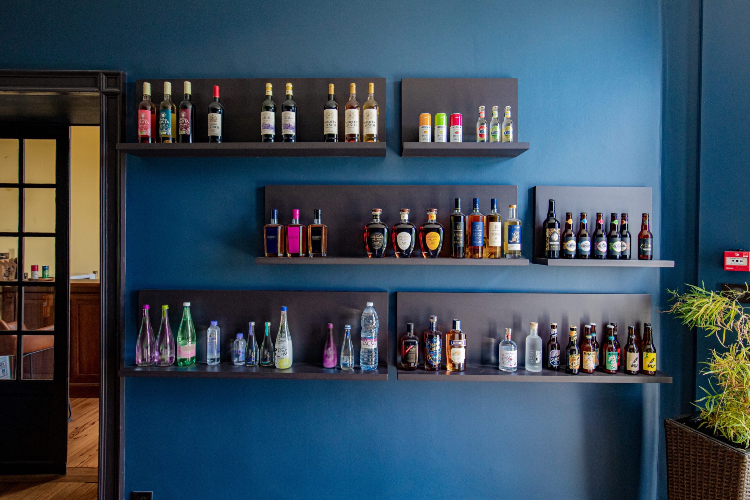 Une s&eacute;lection soigneusement organis&eacute;e de spiritueux et de boissons en bouteille sur d'&eacute;l&eacute;gantes &eacute;tag&egrave;res murales sur un fond bleu saisissant, dispos&eacute;es en &eacute;tapes pour rehausser l'ambiance d'un caf&eacute;-restaurant.