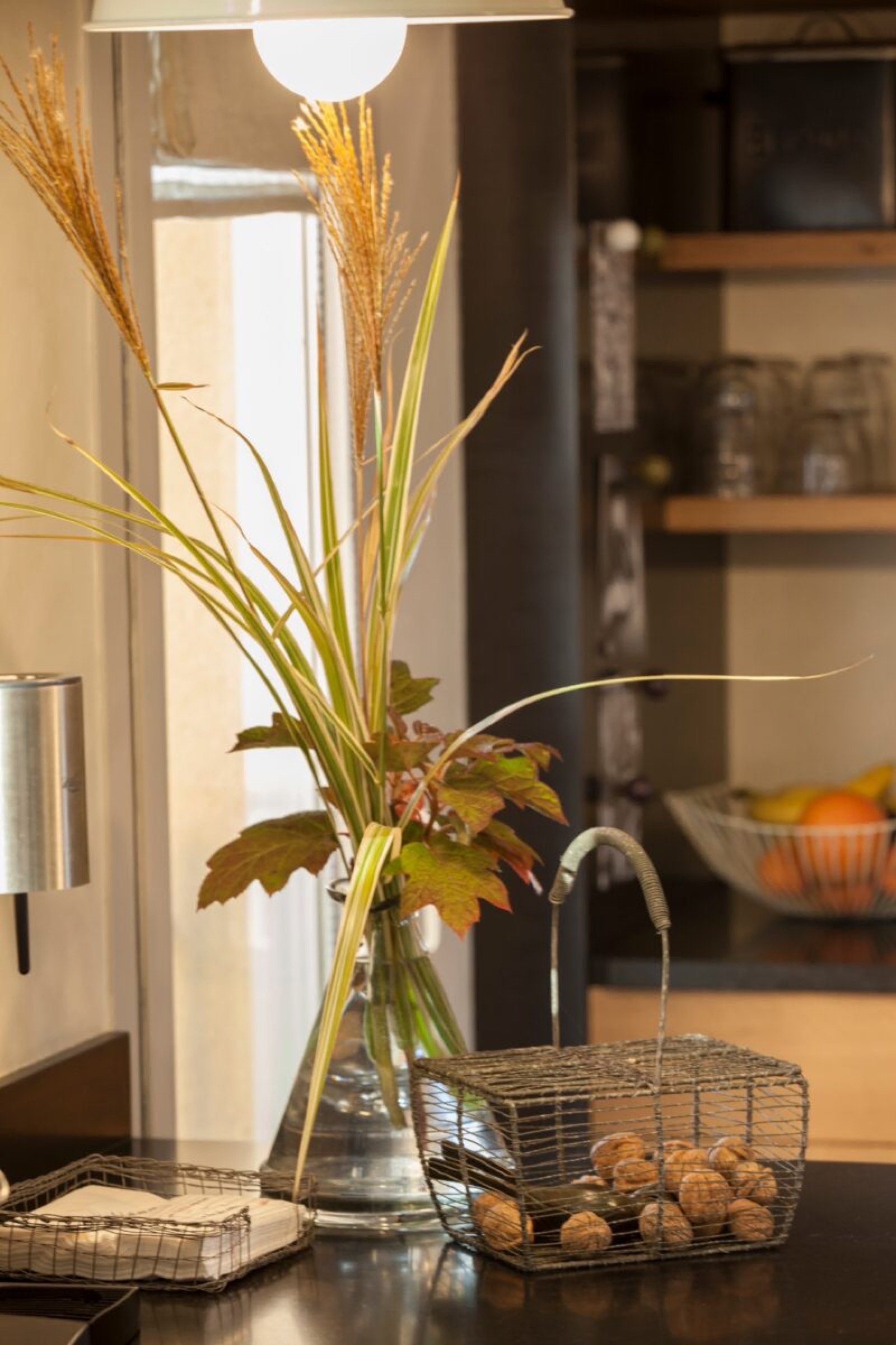 Un coin chaleureux et confortable d'une pi&egrave;ce comprenant un vase avec des herbes et des feuilles s&eacute;ch&eacute;es, un panier avec des noix et une lampe invitante, le tout sur une table en bois sur fond de lumi&egrave;re douce.
