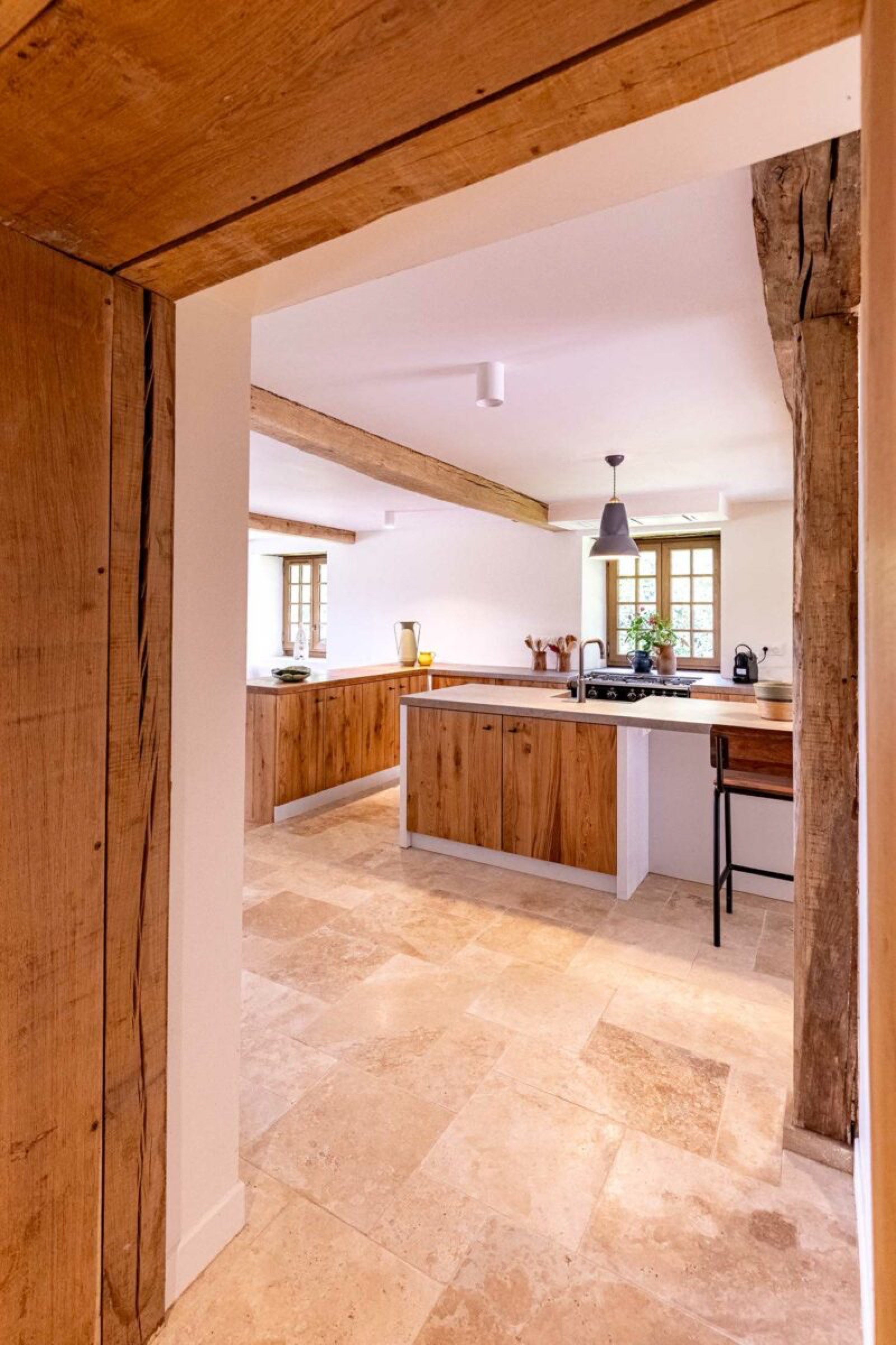 Une cuisine moderne au charme rustique comprend des armoires en bois, des poutres en bois et un sol en carrelage en pierre claire. Il dispose d'un &icirc;lot de cuisine avec &eacute;vier, d'une suspension et de grandes fen&ecirc;tres laissant entrer la lumi&egrave;re naturelle. Un tabouret est plac&eacute; &agrave; c&ocirc;t&eacute; de l&rsquo;&icirc;lot, mettant en valeur une architecture d&rsquo;int&eacute;rieur impeccable.