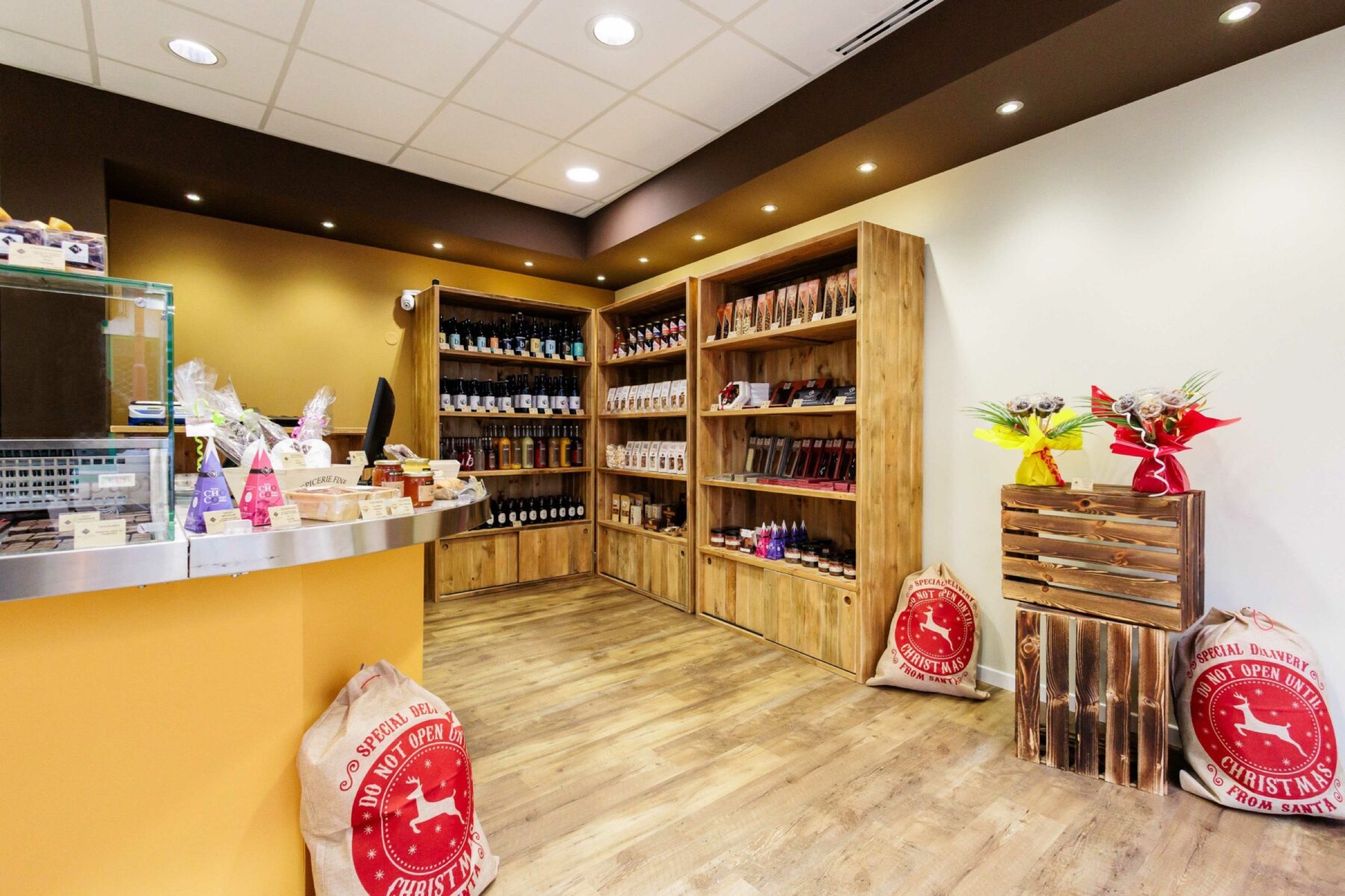 Une chocolaterie cosy avec des étagères en bois présentant divers produits chocolatés. Le comptoir de gauche est doté d'un écran d'ordinateur et est décoré d'articles cadeaux emballés. Le sol est en bois et l’éclairage est chaleureux, créant une atmosphère accueillante, grâce à notre équipe dédiée projets d’agencement intérieur.