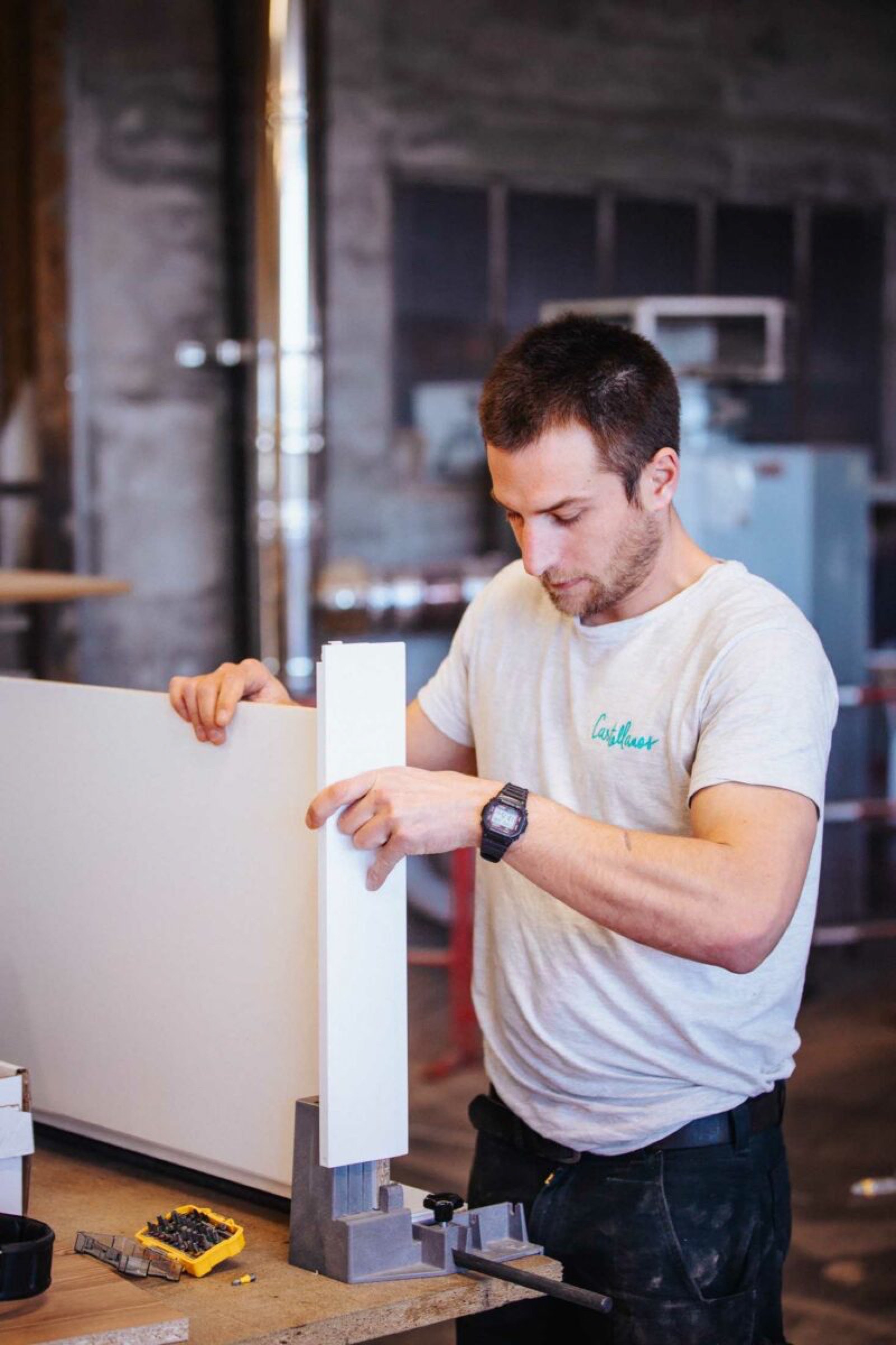 Un homme portant un T-shirt blanc et une montre-bracelet travaille sur un projet de menuiserie. Il assemble un meuble en maintenant deux panneaux blancs ensemble sur un &eacute;tabli. Divers outils et mat&eacute;riaux sont diss&eacute;min&eacute;s autour de l&rsquo;&eacute;tabli, rappelant un entretien de L&eacute;a avec une stagiaire en architecture d&rsquo;int&eacute;rieur.