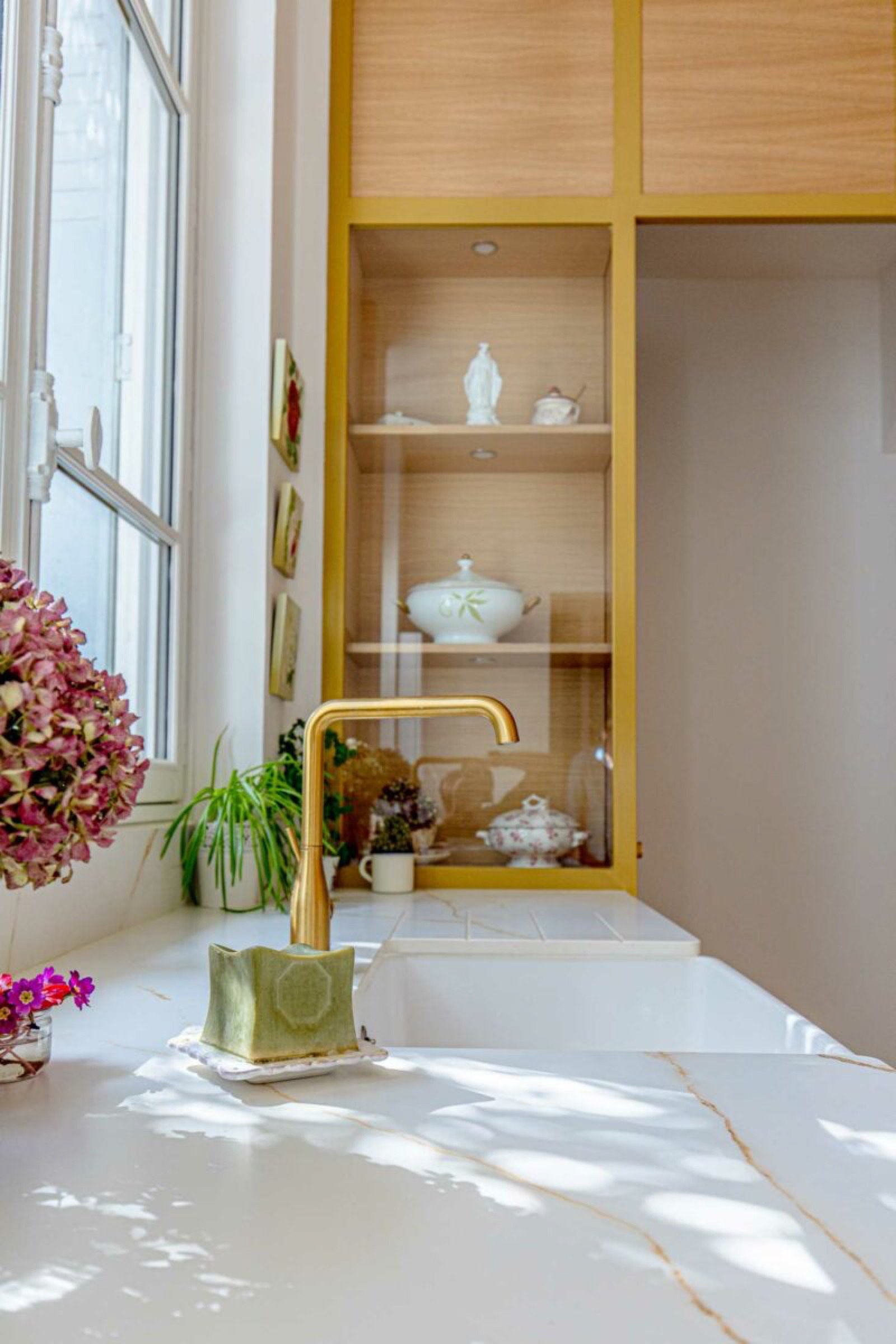Un coin cuisine bien éclairé avec un comptoir en marbre, un robinet doré et une tasse verte en céramique. Une fenêtre sur la gauche laisse entrer la lumière naturelle. Au fond, une armoire en bois avec des étagères en verre présentant des objets décoratifs. Dans le cadre de nos derniers projets d’agencement intérieur, une élégante plante d’hortensia rose orne le comptoir.