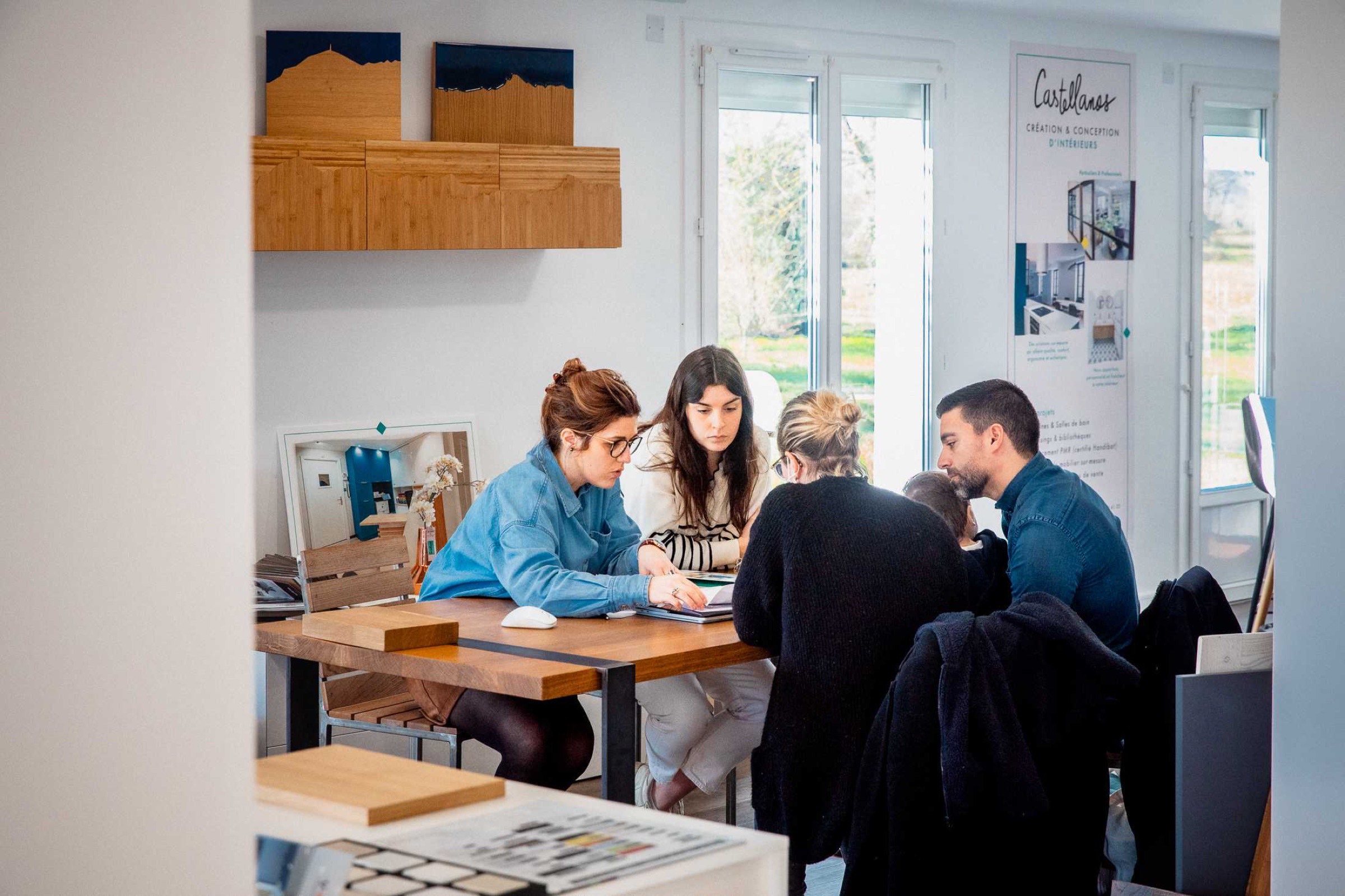 Quatre personnes, dont L&eacute;a, sont assises autour d'une table dans un bureau lumineux. Ils consultent des documents et discutent intens&eacute;ment, peut-&ecirc;tre pour un entretien sur l&rsquo;architecture d&rsquo;int&eacute;rieur. Le groupe semble concentr&eacute; et engag&eacute;. Le bureau dispose de grandes fen&ecirc;tres, d&rsquo;&oelig;uvres d&rsquo;art sur des &eacute;tag&egrave;res et d&rsquo;une banni&egrave;re en arri&egrave;re-plan.