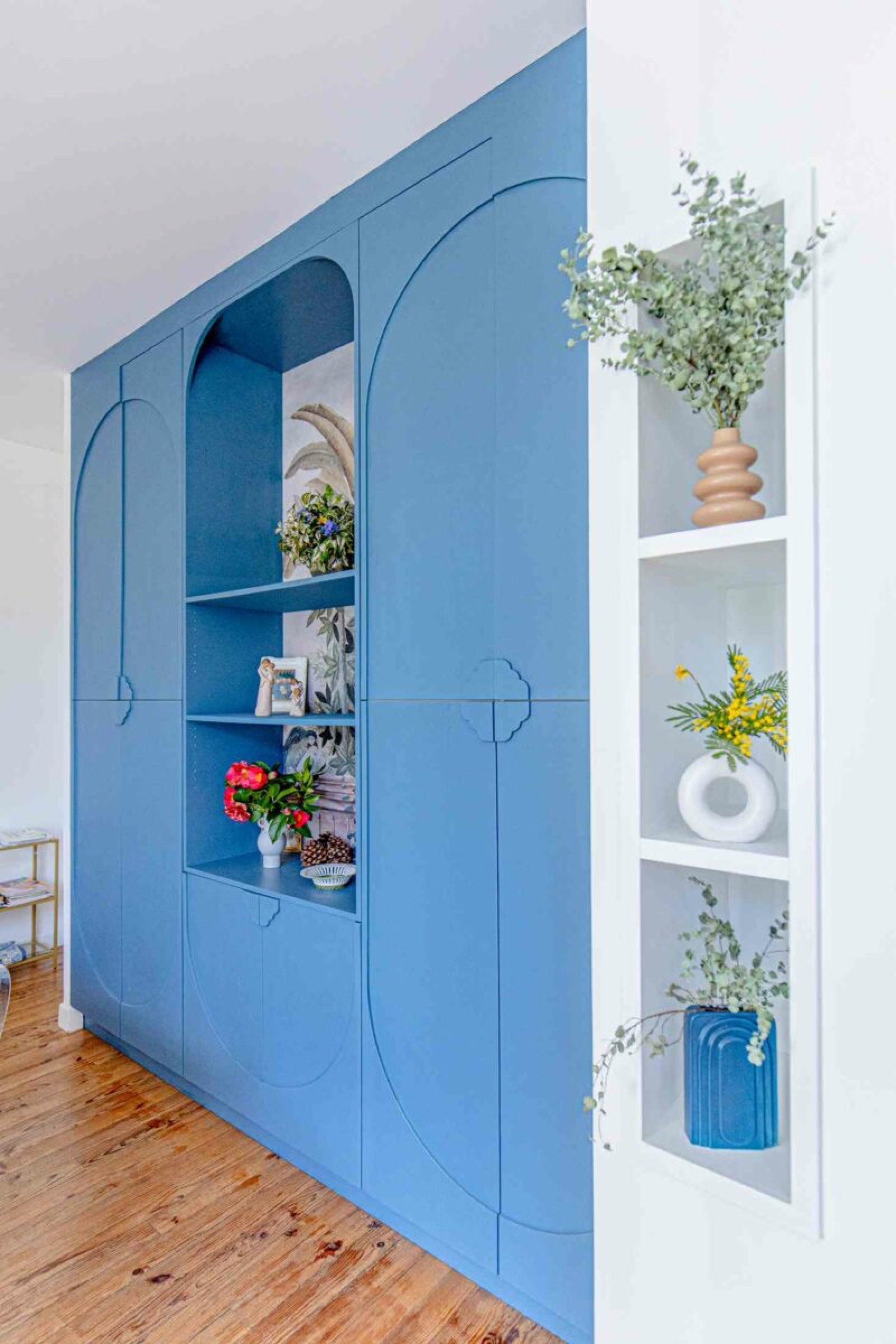 Une bibliothèque intégrée bleue au design moderne, dotée de poignées incurvées découpées, s'étend du sol au plafond. Il contient un mélange de livres, de photos encadrées et d'objets de décoration, notamment des fleurs. À côté, une petite étagère niche blanche créée par notre équipe affiche davantage de décoration et de plantes – un projet d’agencement intérieur idéal.