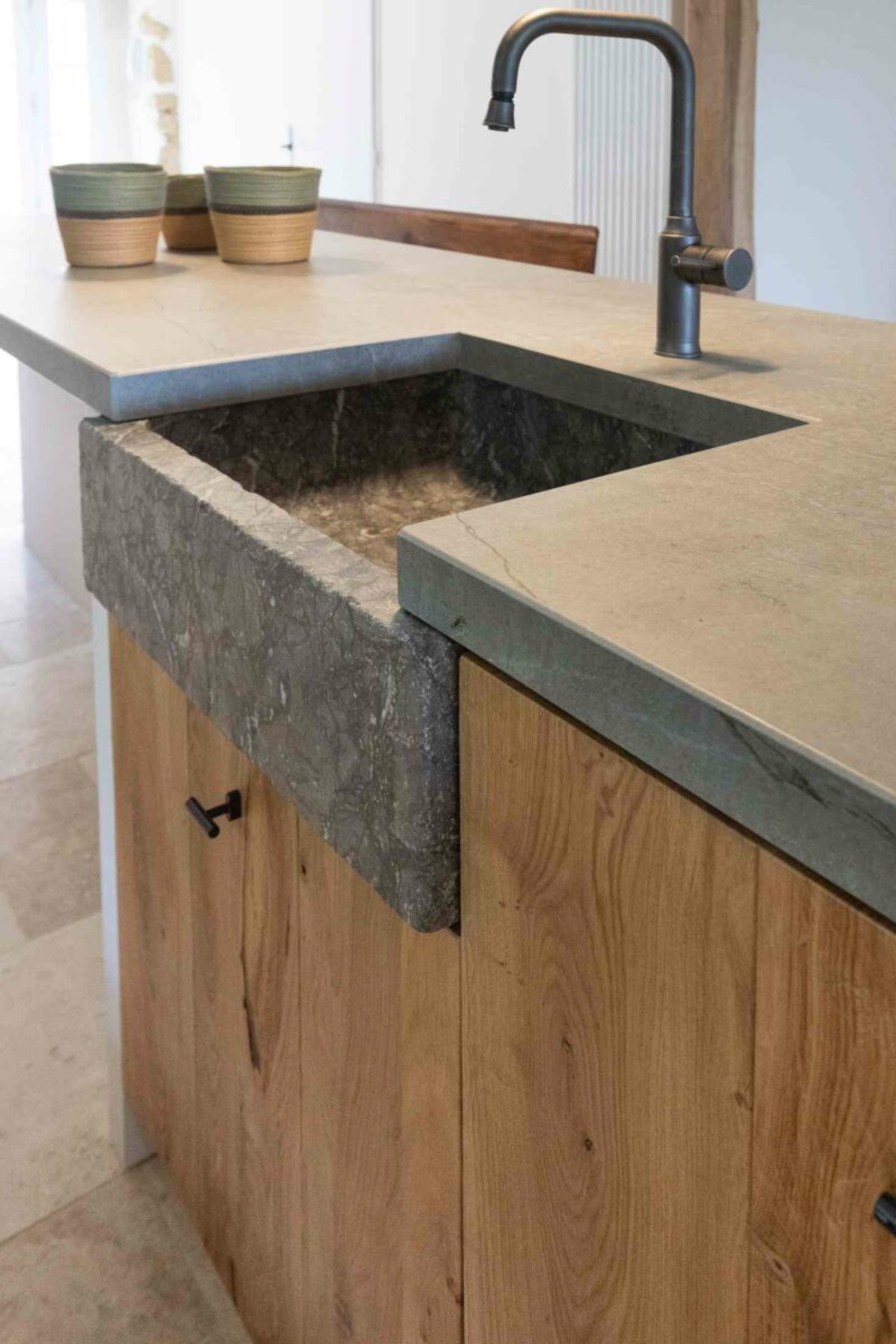 Une cuisine aux armoires en bois pr&eacute;sente un &eacute;vier en pierre rustique encastr&eacute; dans un comptoir beige. Deux petits bols verts sont plac&eacute;s sur le comptoir pr&egrave;s du robinet moderne et minimaliste. L'esth&eacute;tique g&eacute;n&eacute;rale est terreuse et naturelle, s'alignant parfaitement avec les tendances d&eacute;co de l'am&eacute;nagement 2025.