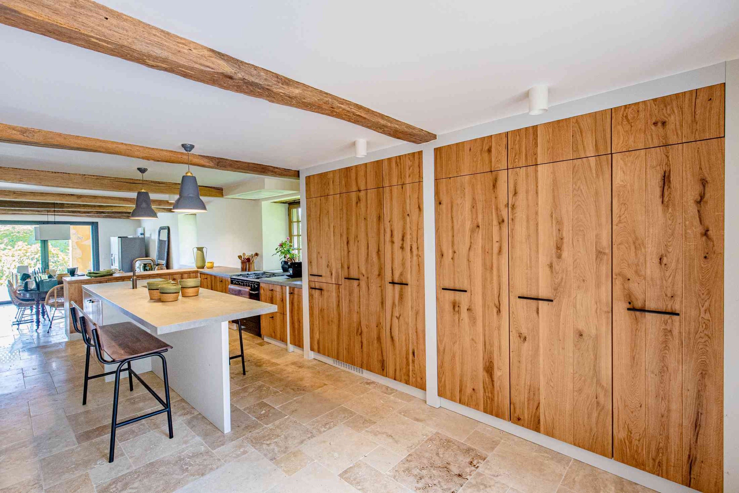 Une cuisine moderne avec des meubles en bois, un &icirc;lot avec un comptoir blanc et des tabourets de bar. Des poutres en bois apparentes sont au plafond et de grandes fen&ecirc;tres offrent une abondante lumi&egrave;re naturelle. En accord avec les tendances d&eacute;co 2025, la cuisine se prolonge par un coin repas avec un am&eacute;nagement bien pens&eacute;.