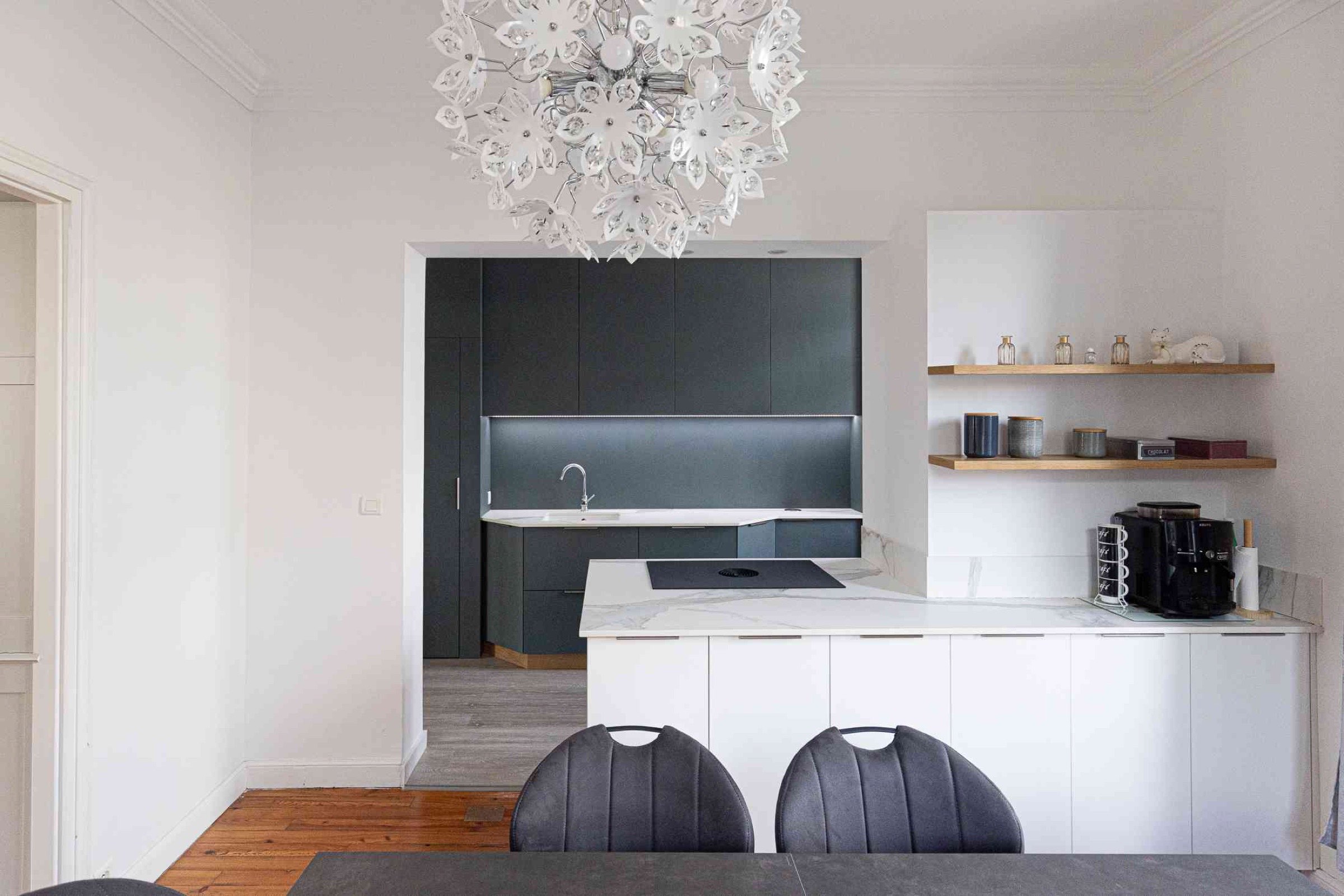 Cuisine minimaliste avec des armoires sombres, un comptoir &eacute;l&eacute;gant et des murs blancs. Un lustre moderne au motif floral est suspendu au plafond. Adoptant les tendances d&eacute;co 2025, la pi&egrave;ce comprend des &eacute;tag&egrave;res ouvertes avec divers objets et une cafeti&egrave;re. Une table &agrave; manger et des chaises sont au premier plan.
