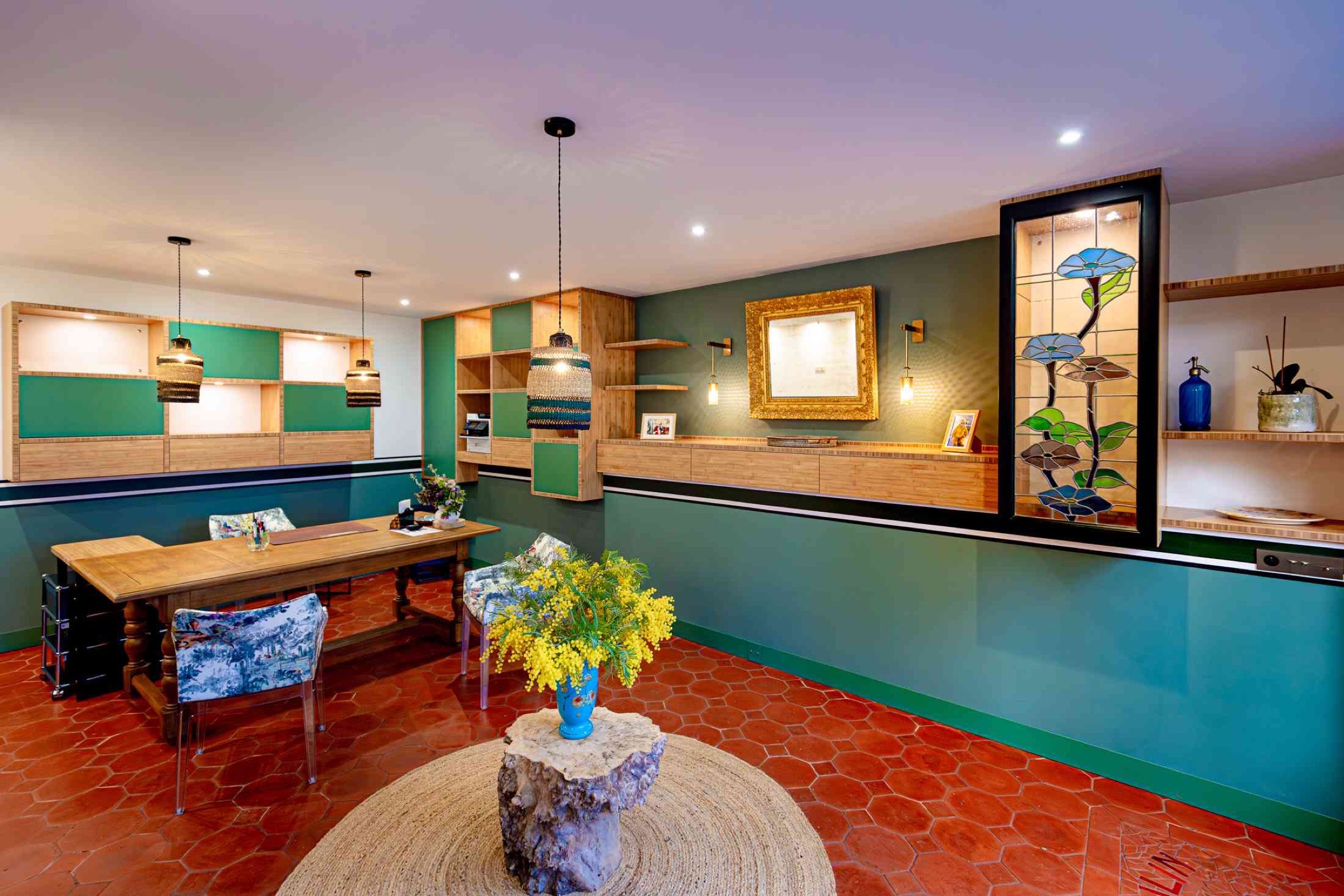 Un bureau moderne avec un mur d'accent vert, une table en bois et des chaises rembourr&eacute;es qui suivent les derni&egrave;res tendances en mati&egrave;re de d&eacute;coration. La pi&egrave;ce dispose d'un sol carrel&eacute; rouge, de luminaires suspendus, d'&eacute;tag&egrave;res d&eacute;coratives et d'un grand vitrail ornemental repr&eacute;sentant des fleurs bleues. Un vase avec des fleurs jaunes est pos&eacute; sur la table.