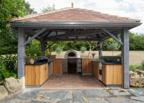 Un charmant pavillon de cuisine extérieure comprenant un four à pizza, un grill et des armoires en bois conçus par un cuisiniste qualifié, niché dans un jardin verdoyant avec une allée en pierre qui y mène.