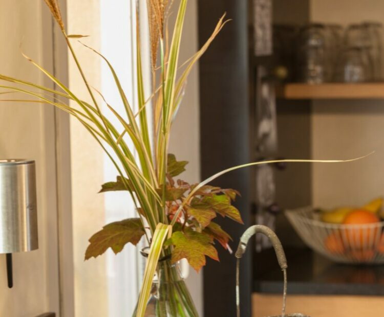 Un coin chaleureux et confortable d'une pièce comprenant un vase avec des herbes et des feuilles séchées, un panier avec des noix et une lampe invitante, le tout sur une table en bois sur fond de lumière douce.