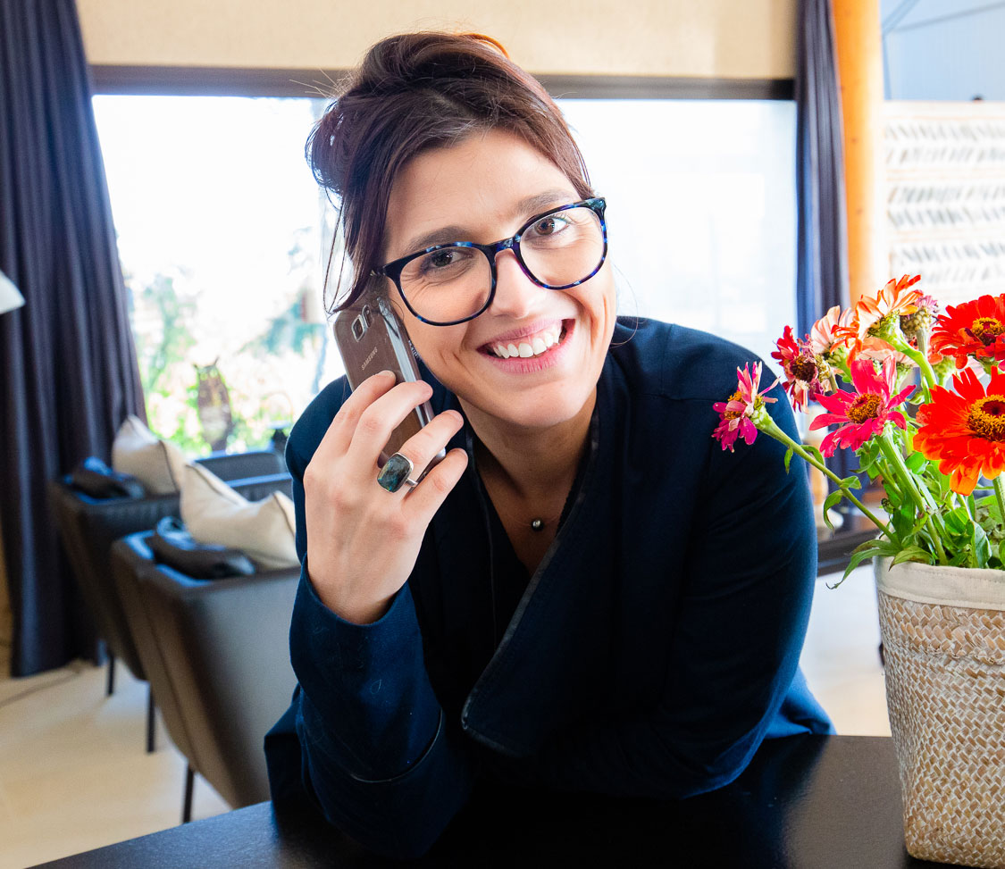 Une femme joyeuse portant des lunettes sourit tout en discutant sur son téléphone portable, assise confortablement à l'intérieur avec un bouquet de fleurs aux couleurs vives à proximité.