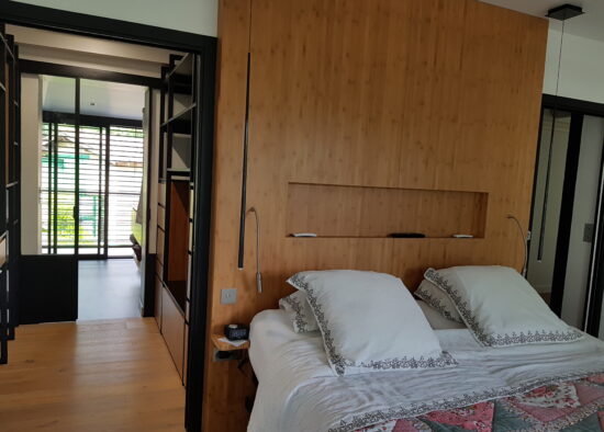 Une chambre confortable et moderne dotée d'un grand lit avec une couette décorative, de murs recouverts de bambou, de lampes de lecture et d'une vue sur une pièce attenante avec lumière naturelle filtrant à travers les stores. De plus, l'espace dispose