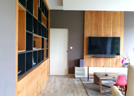 Un salon moderne avec un mélange de textures bois, conçu sur mesure par un architecte d'intérieur à Tarbes, comprenant une grande bibliothèque vide, une télévision murale et un décor minimaliste.