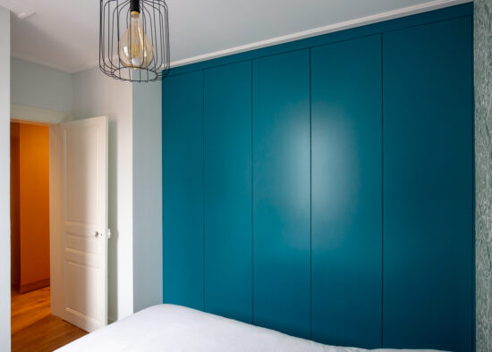 Une chambre moderne avec un mur d'accent bleu sarcelle audacieux et une élégante suspension en fil noir, avec une porte contrastée aux tons chauds et des meubles sur mesure par un cuisiniste à Tarbes en arrière-plan.