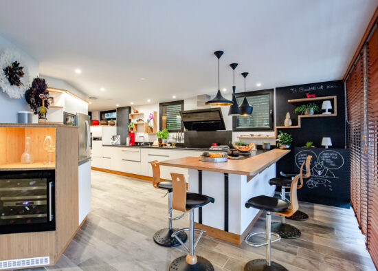 Une cuisine moderne avec des armoires blanches et un îlot central doté de tabourets de bar, complétée par un mur en tableau et un éclairage chaleureux, conçue par un cuisiniste professionnel à Pau.
