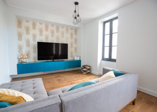 Un salon moderne au design minimaliste comprenant un canapé gris, une console multimédia bleu vif, des meubles sur mesure comprenant une télévision à écran plat sur un mur à motifs et une suspension géométrique.