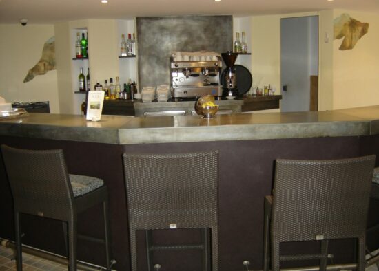 Un comptoir de café moderne avec des machines à expresso, des tabourets de bar et une sélection personnalisée de boissons sur les étagères, dégageant une atmosphère chaleureuse et accueillante.