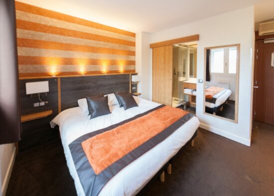 Une chambre d'hôtel moderne et cosy avec un grand lit, des touches orange, des murs aux motifs rayés et une armoire à glace reflétant une partie supplémentaire de la chambre conçue par un cuisiniste à Tarbes.