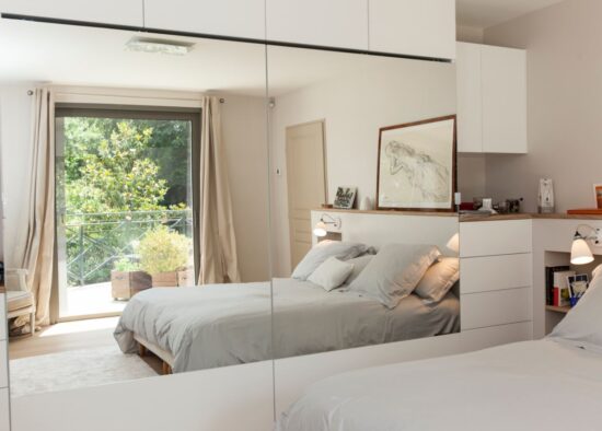 Une chambre moderne avec un grand miroir reflétant le lit et une fenêtre donnant sur l'extérieur, créant une ambiance aérée et spacieuse. L'inclusion de meubles sur mesure ajoute à l'élégance de la pièce