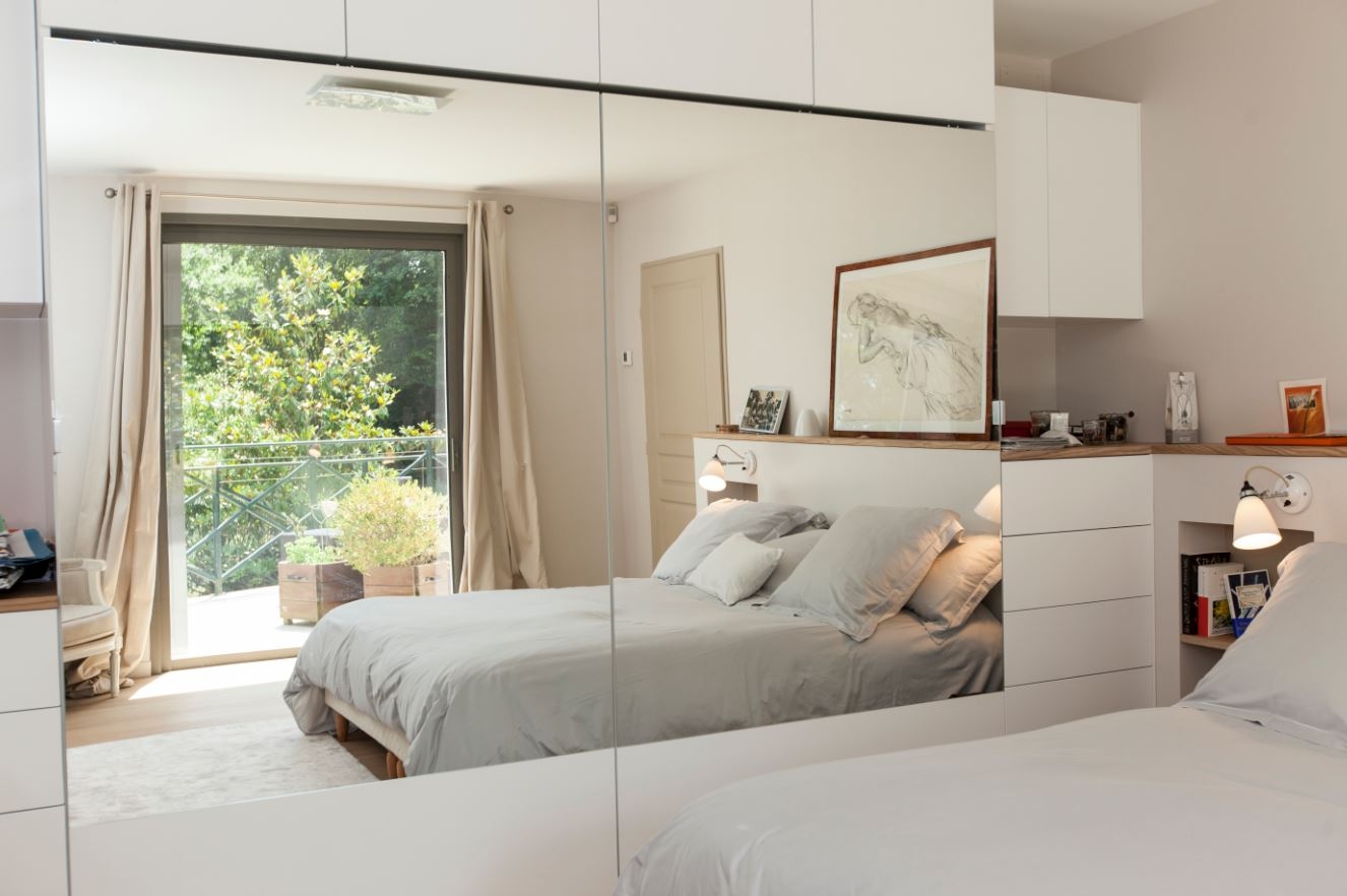 Chambre élégante dotée d'un grand miroir reflétant un lit bien rangé avec une literie grise et d'un balcon laissant entrer la lumière naturelle, créant un espace lumineux et aéré.