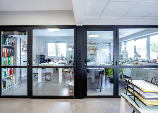 Un espace de bureau lumineux et moderne vu à travers des portes vitrées, comprenant une bibliothèque organisée à gauche, des bureaux avec ordinateurs et paperasse, et des plantes apportant une touche de verdure, conçu par un sur mesure