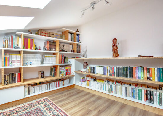 Une chambre mansardée confortable avec des plafonds mansardés, dotée d'étagères sur mesure bien organisées pleines de livres colorés, d'un tapis décoratif sur le parquet et d'un éclairage lumineux.