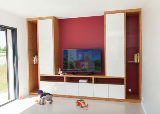 Salon moderne avec un mur d'accent rouge foncé doté d'une télévision à écran plat et d'armoires blanches, avec un enfant jouant par terre près d'une porte vitrée ouverte offrant une vue sur l'extérieur. Le