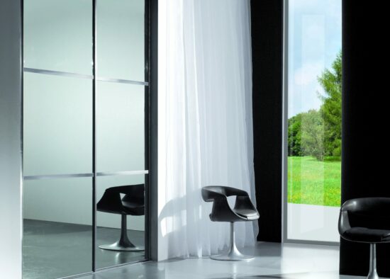 Une chambre moderne dotée de chaises noires élégantes, d'une grande porte vitrée menant à une vue extérieure verdoyante et de rideaux blancs ondulants se balançant doucement dans l'espace aéré et lumineux. L'ajout d'une cuisine