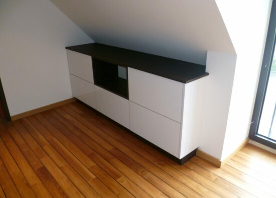 Un buffet blanc moderne avec un plateau noir, conçu comme meubles sur mesure, est placé contre le mur incliné d'une pièce mansardée, complétant le parquet chaleureux et la lumière naturelle.